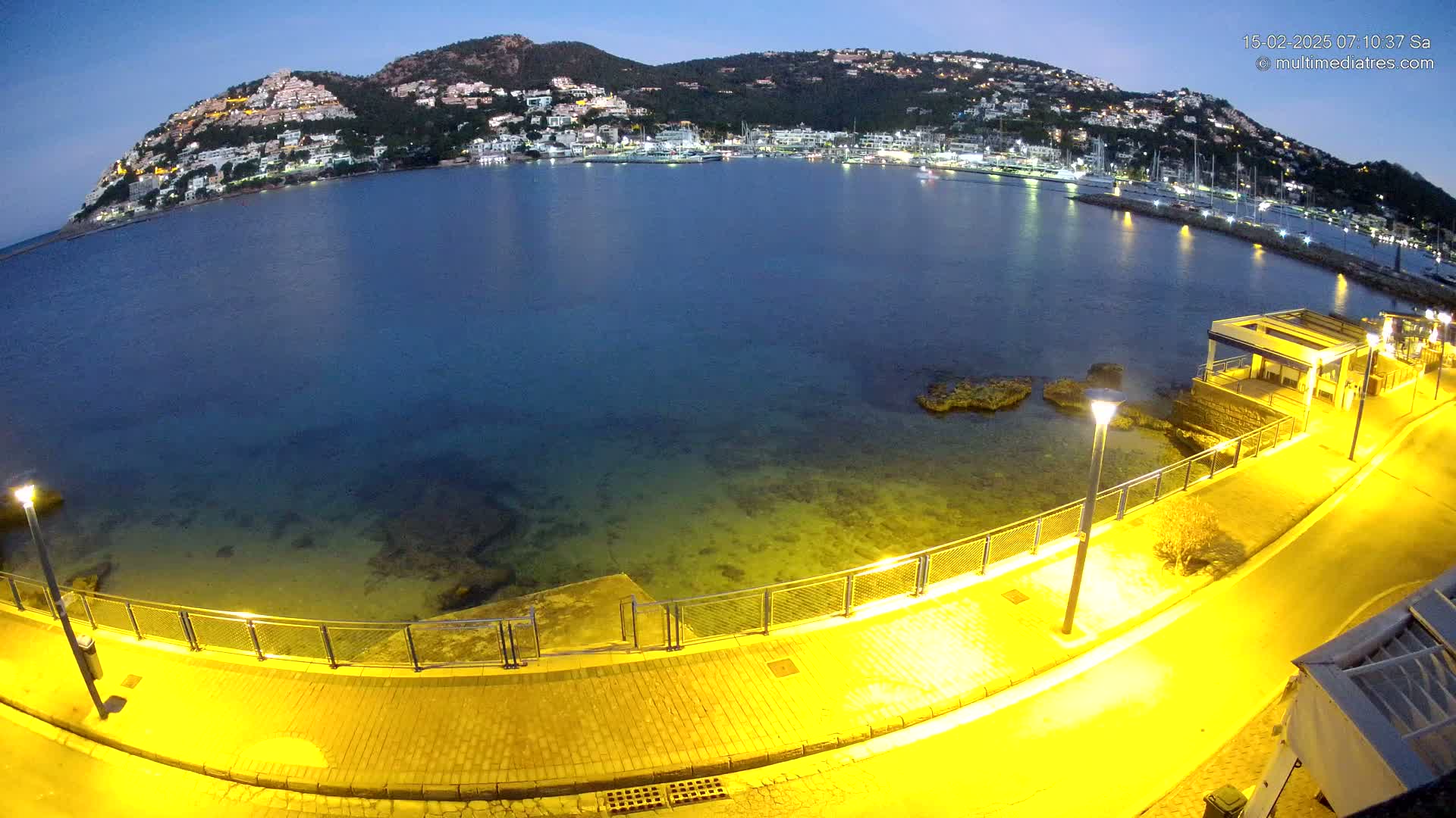 A nighttime view of a paved waterfront walkway illuminated by yellow lights, overlooking a calm, dark-blue sea with a hilltop town in the background.