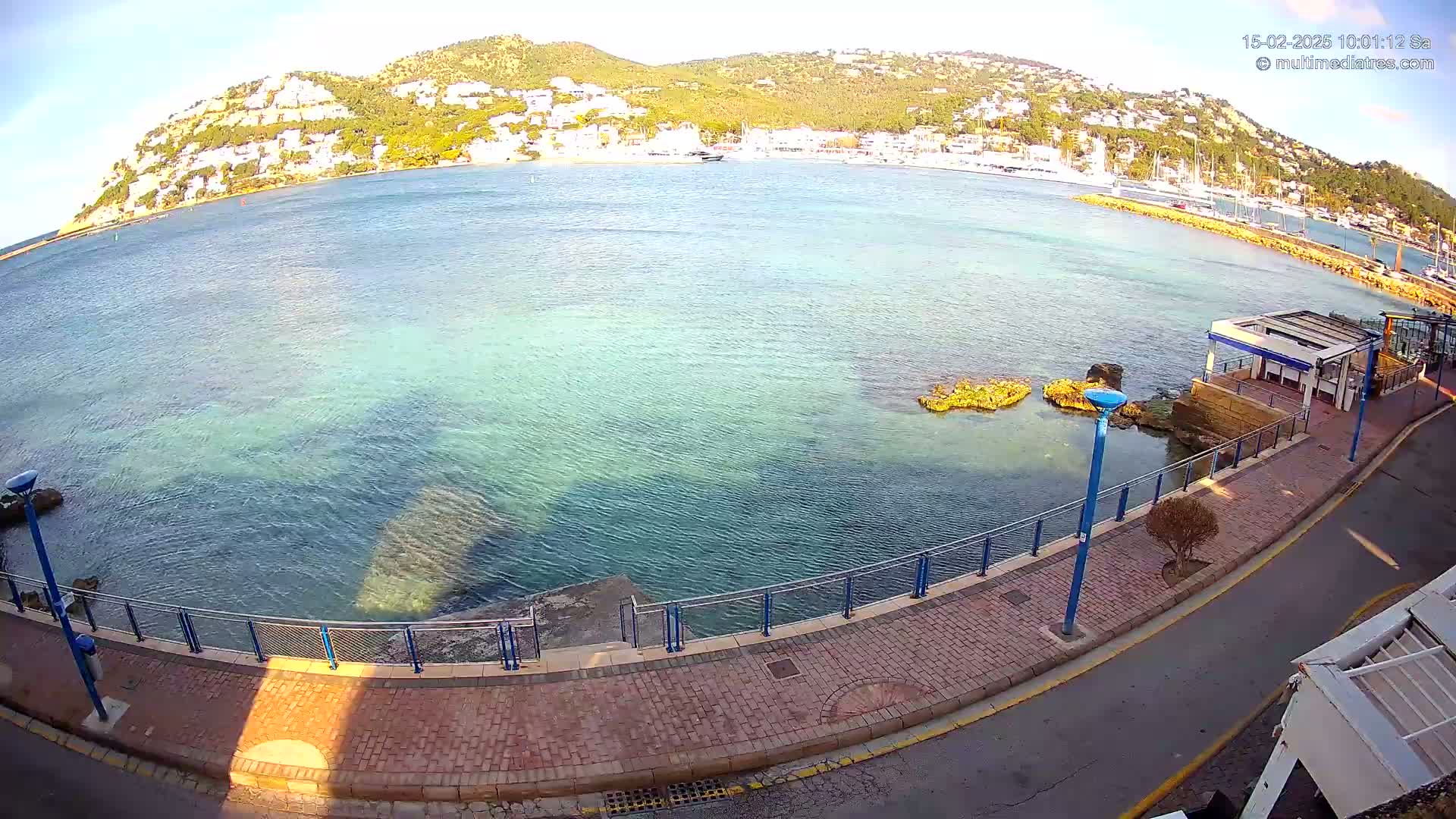 A clear, sunny day overlooks a calm, turquoise bay with a hillside town in the background, seen from a paved waterfront walkway.