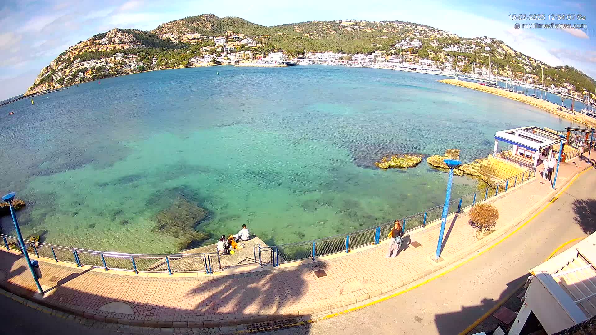A clear, sunny day shows a waterfront scene with calm, shallow, teal-colored water lapping against a paved walkway where several people are relaxing and strolling.