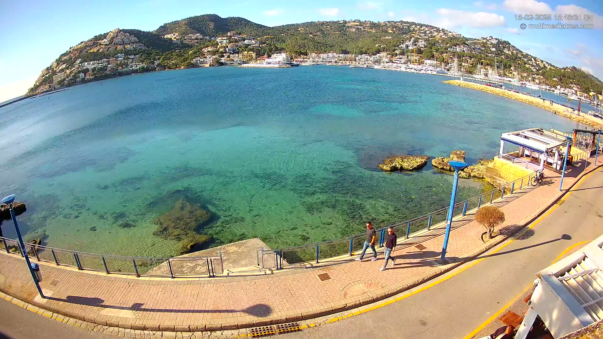 A clear, sunny day overlooks a calm, shallow, turquoise bay with a town nestled on a hillside in the background, and two people walking along a paved waterfront path.
