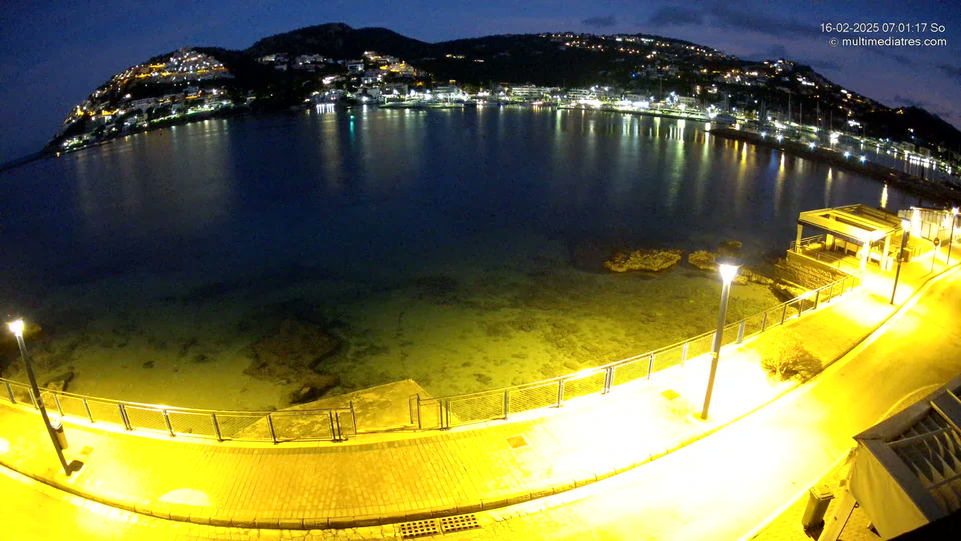 A nighttime, illuminated view of a calm bay with a hilltop town across the water, a paved waterfront path, and a small structure.