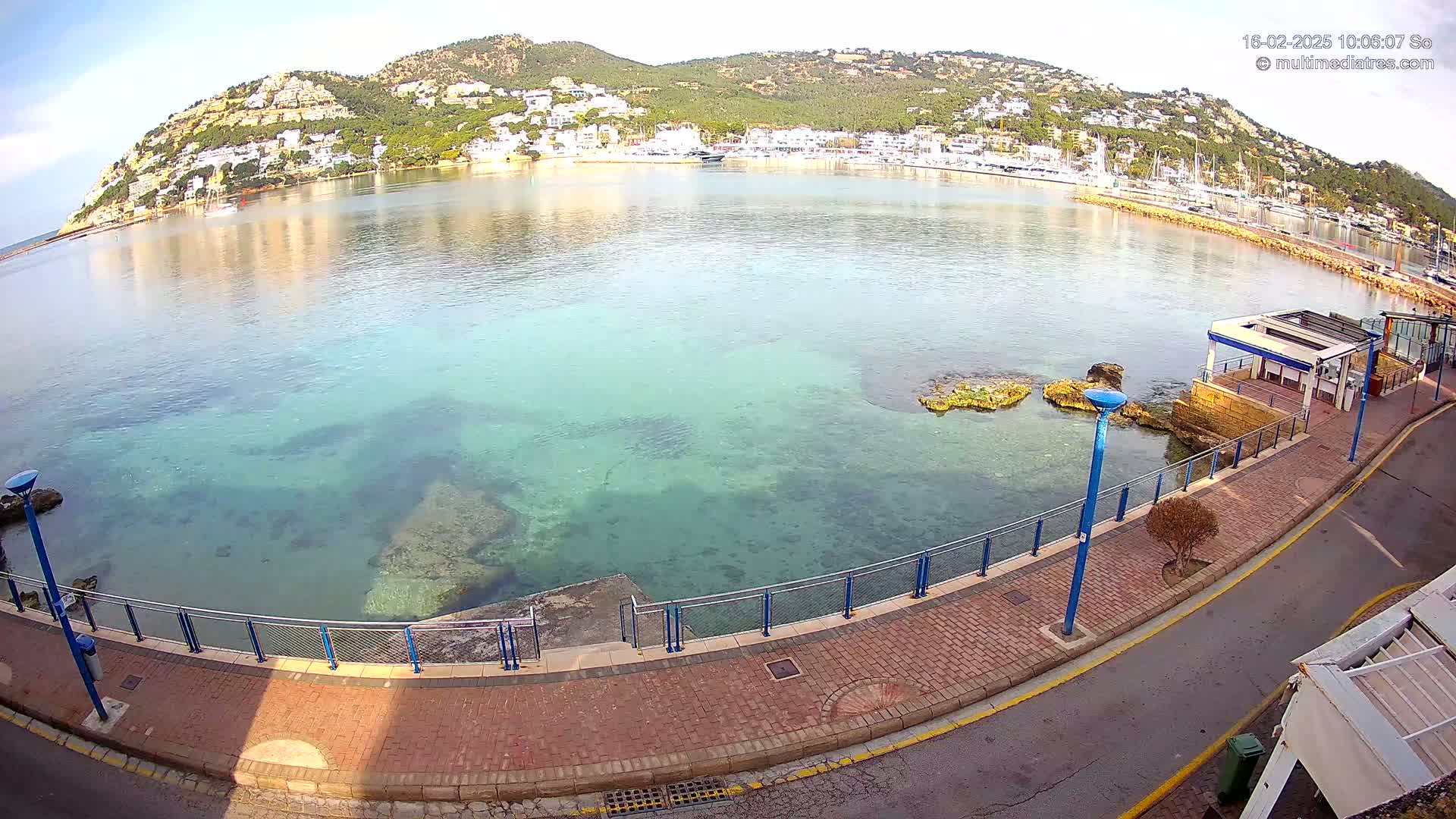 A clear, sunny day shows a calm bay with clear, shallow turquoise water, a paved walkway along the shore, and a hillside town in the background.
