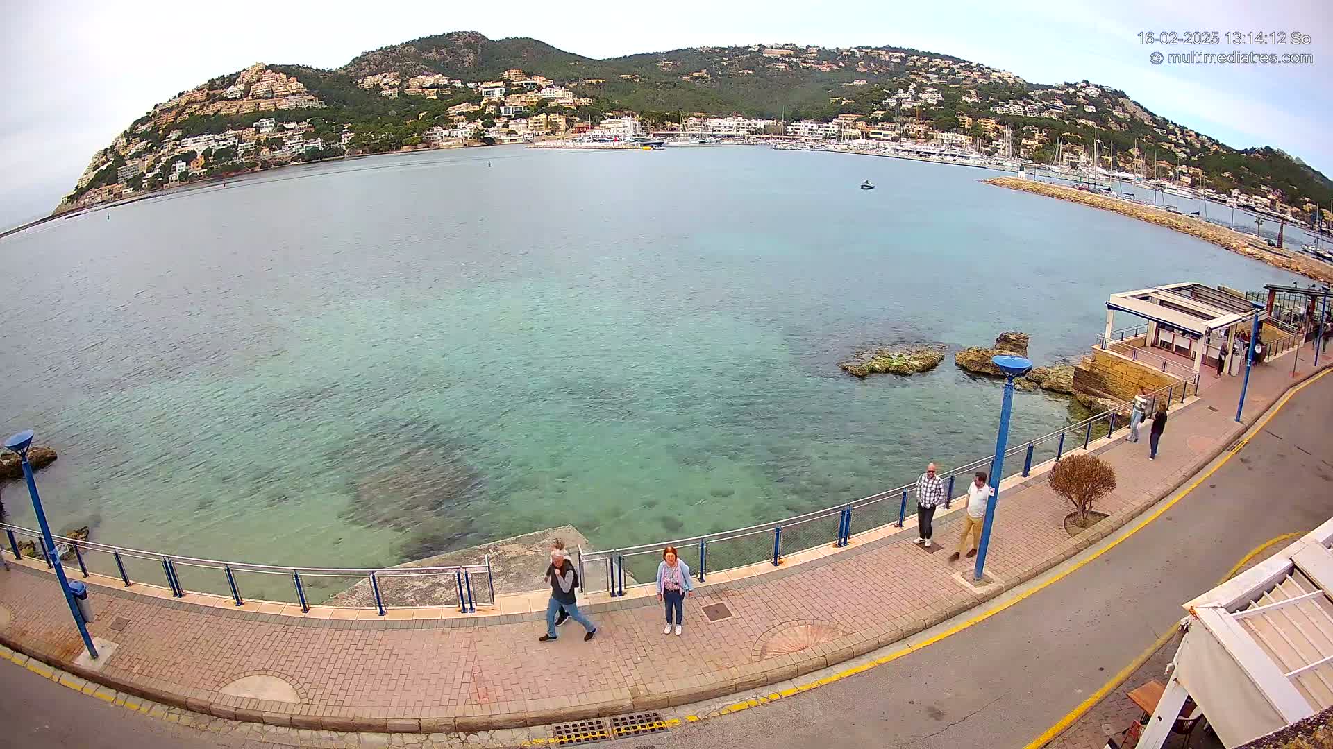 A calm, clear body of water laps a paved walkway lined with people, all overlooking a harbor filled with boats and a hillside town under a mostly cloudy sky.