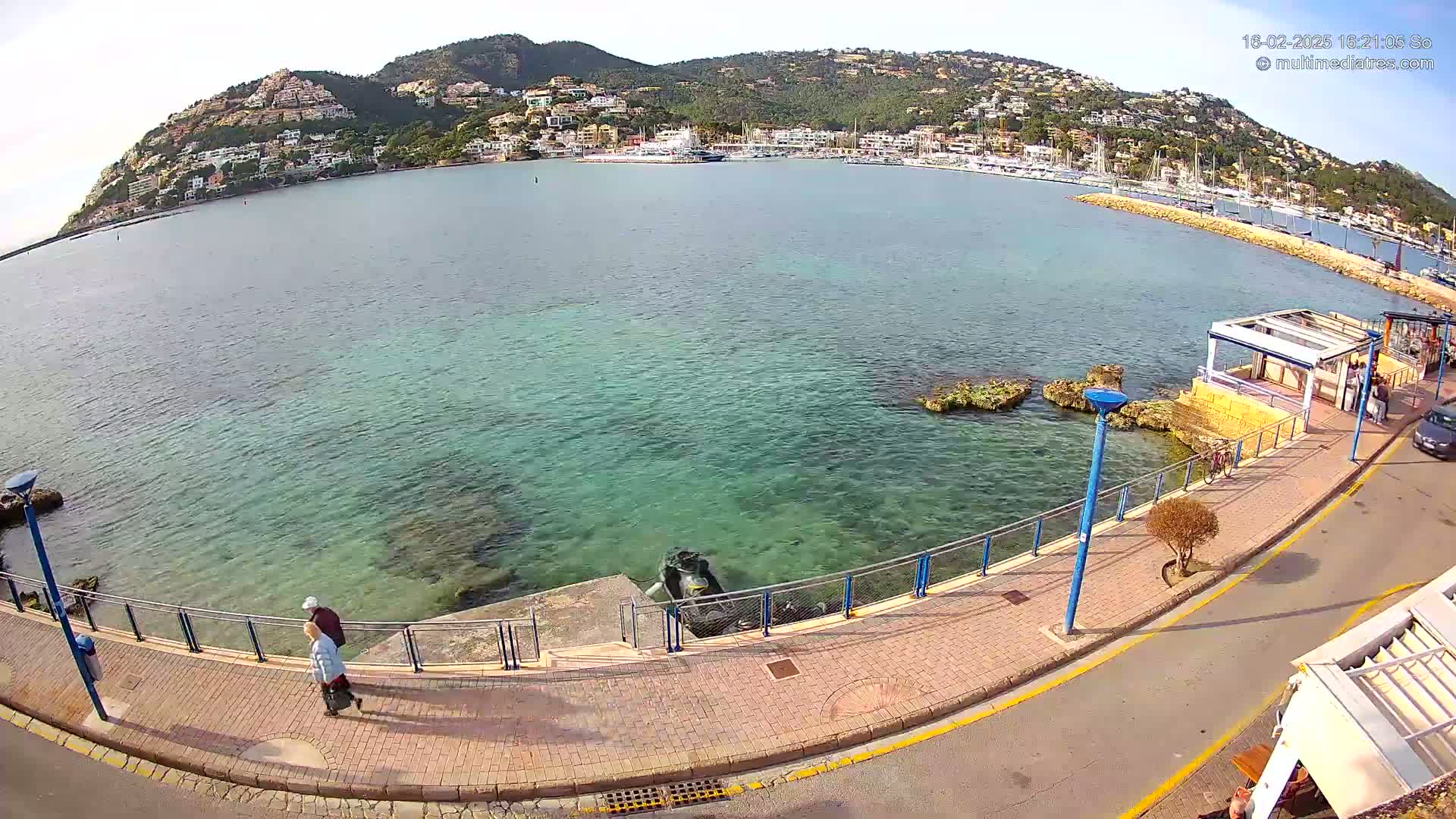 A clear, sunny day overlooks a calm, teal-colored bay with a town nestled on a hillside in the background, a paved walkway along the water's edge, and two people walking by a railing.