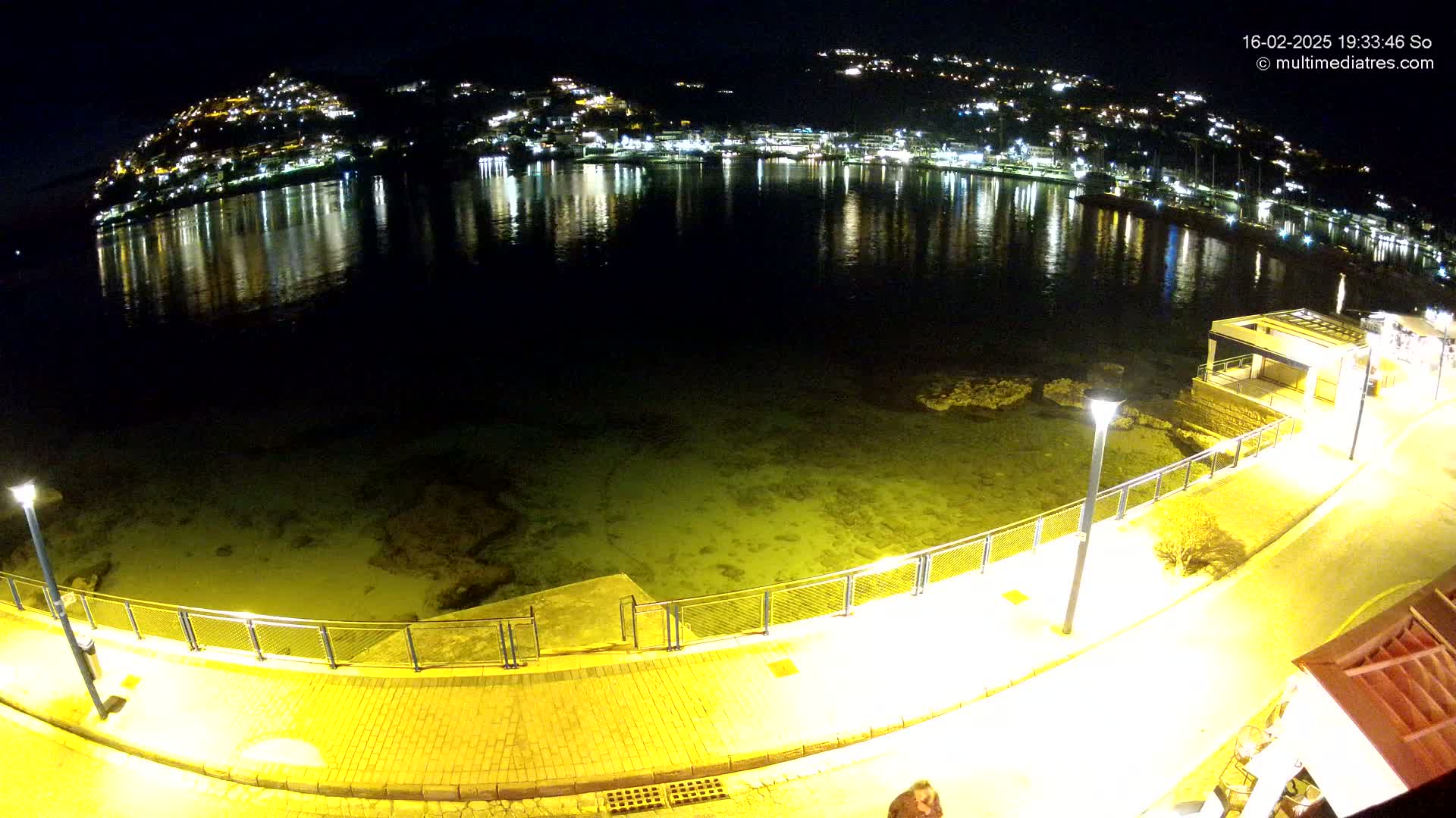 A nighttime view of a waterfront promenade with a calm body of water reflecting the lights of a hilltop town.