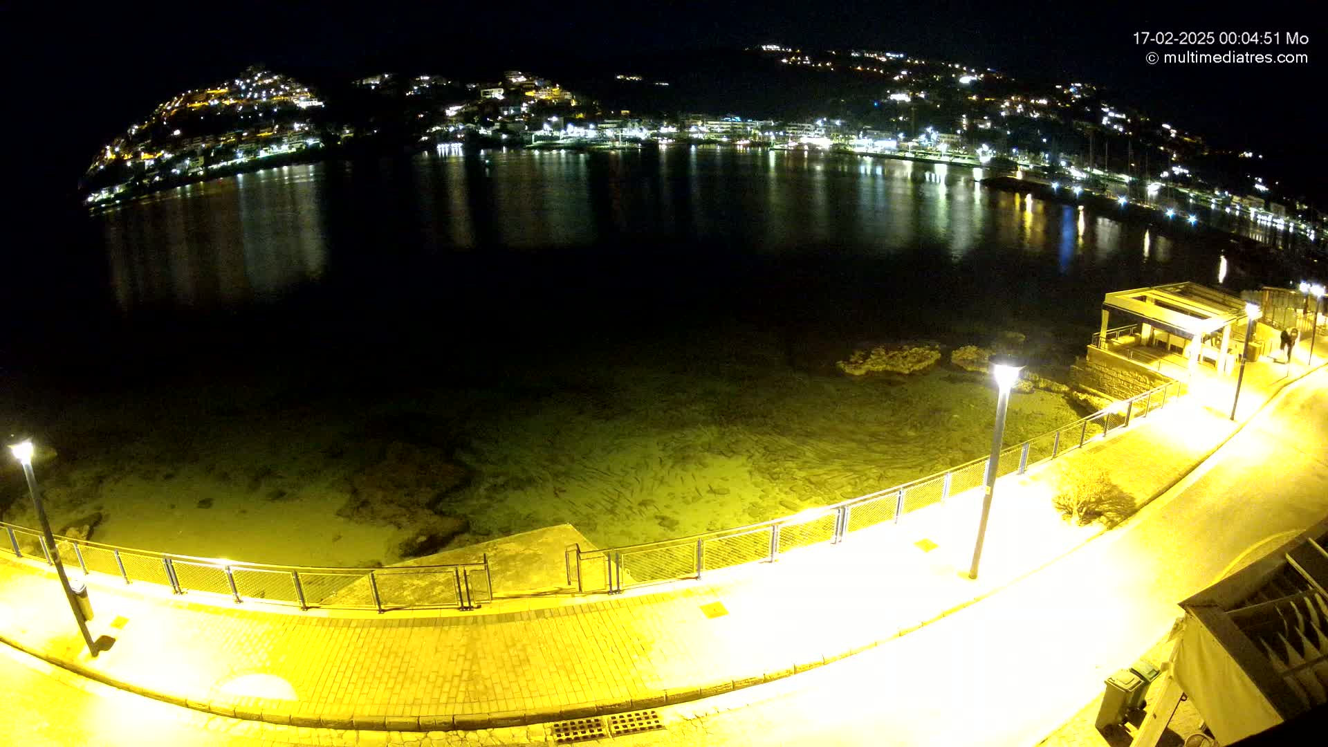 A nighttime view of a calm, shallow body of water reflecting the lights of a town nestled on a hillside, with a paved walkway and lighting along a waterfront.