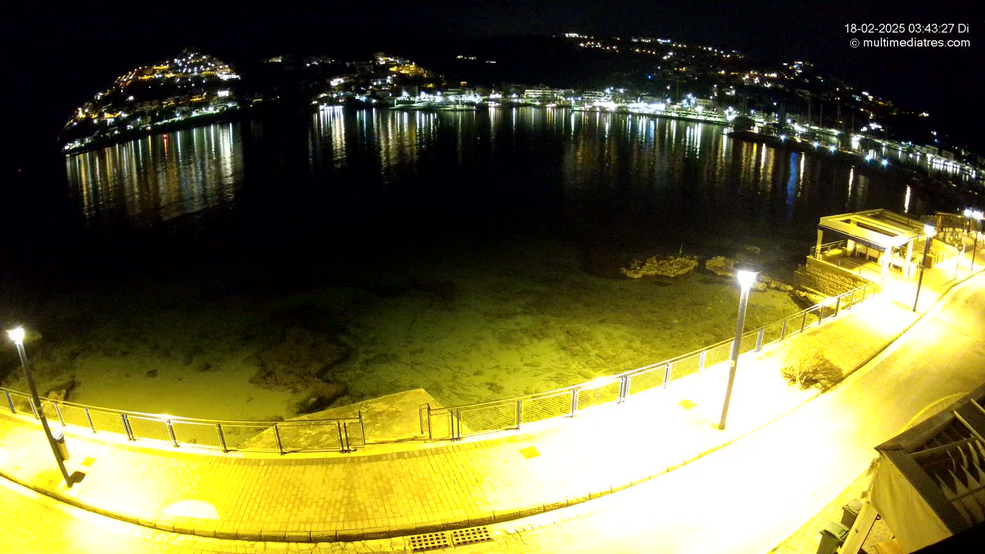 A nighttime view of a waterfront promenade with calm water reflecting the lights of a town on a hill across the bay.