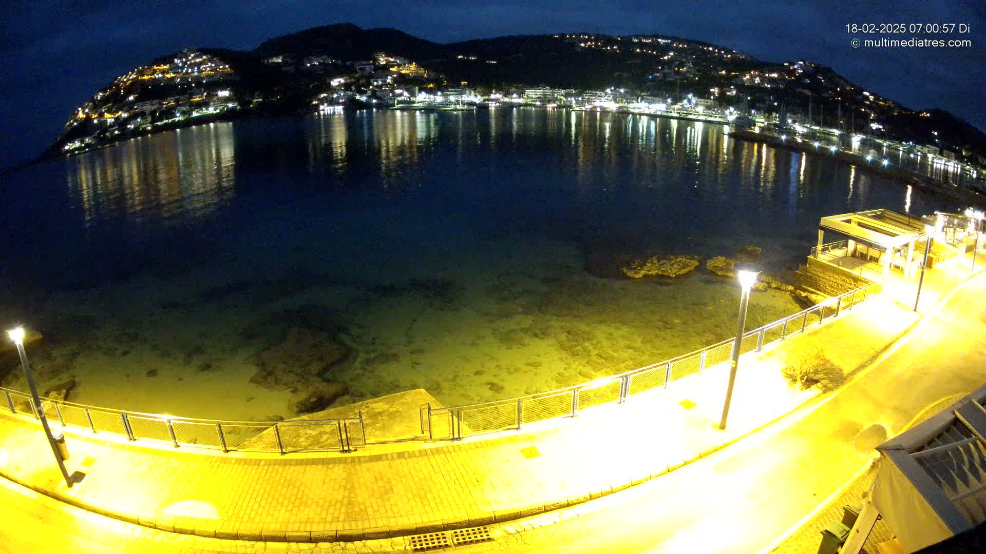 A nighttime view of a calm bay reflecting the lights of a hillside town, with a brightly lit walkway curving along the waterfront.