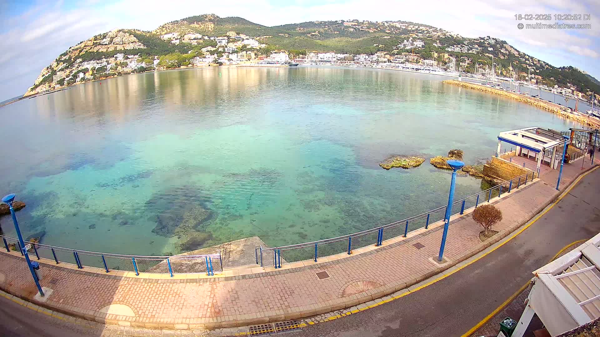 A clear, calm bay with shallow, clear turquoise water laps a paved waterfront walkway next to a small building, all nestled below a hillside town on a sunny day.