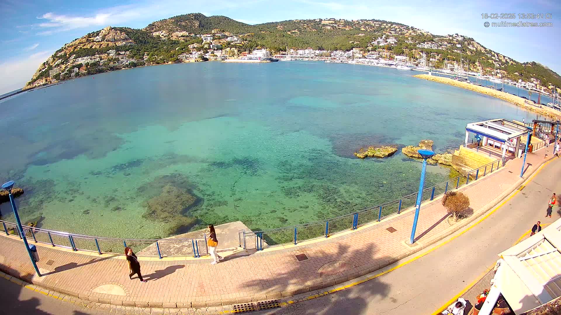 A clear, sunny day reveals a harbor filled with boats, a hill dotted with buildings, and a seaside promenade with a few pedestrians, all overlooking a calm, shallow, clear turquoise sea.