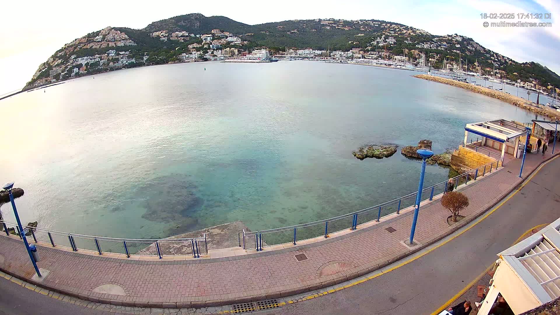 A calm, clear body of water laps against a paved waterfront walkway next to a hillside town under a bright, overcast sky.