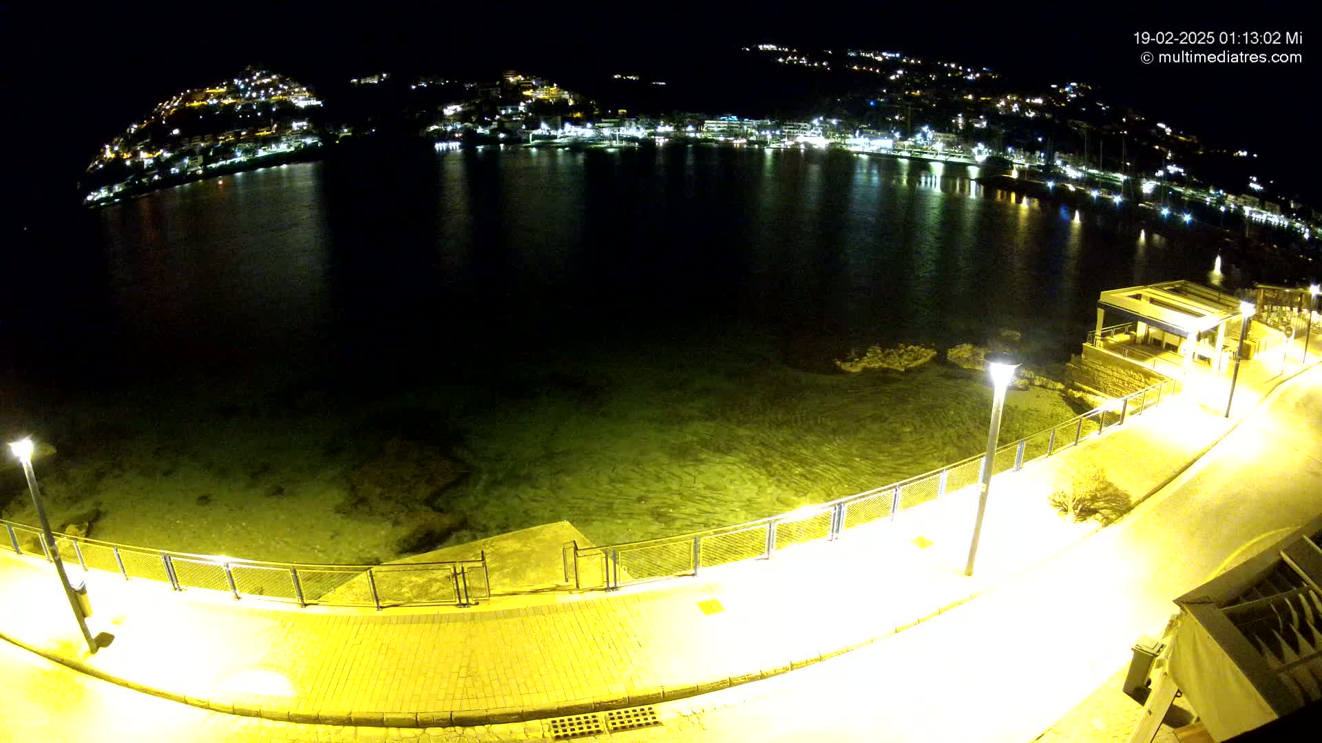 A nighttime view of a calm, dark-green body of water illuminated by lights from a town on a hill in the background, with a paved walkway and lampposts along the water's edge.