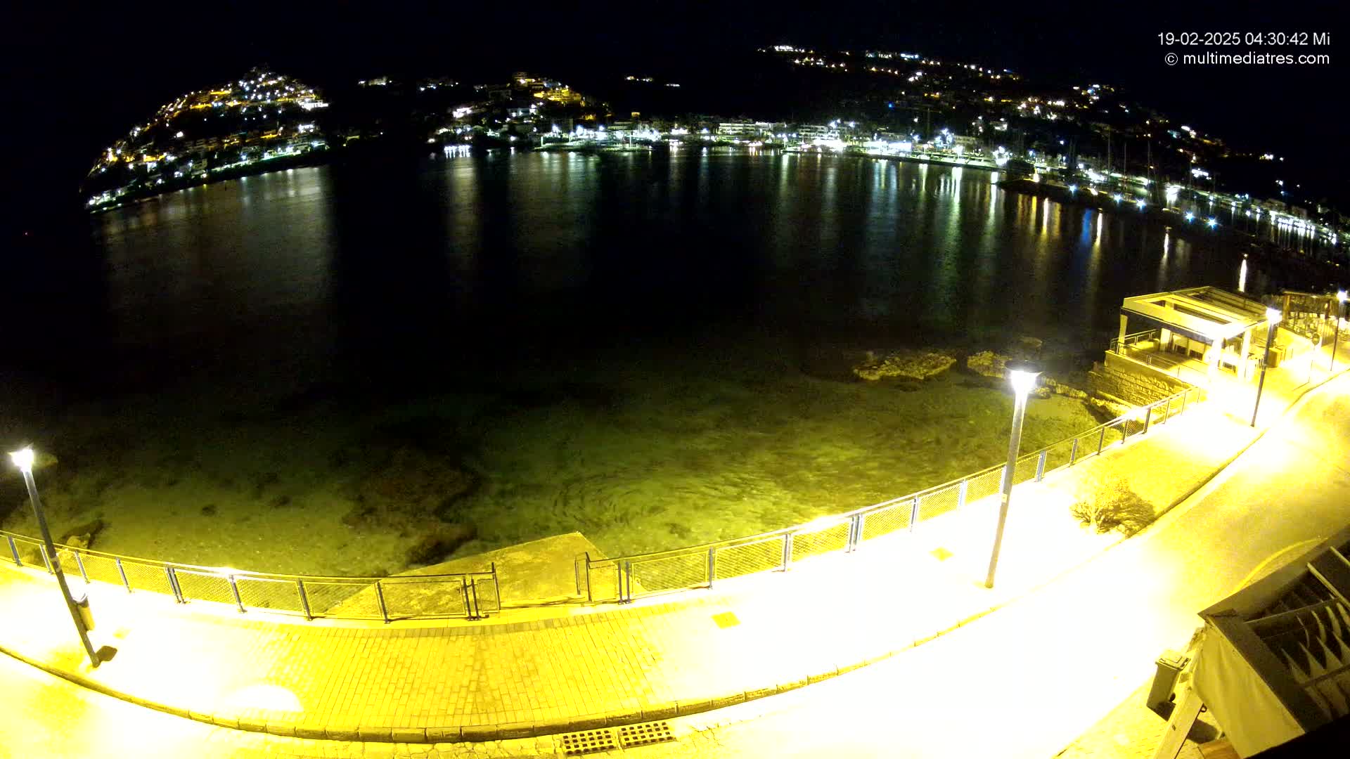 A nighttime, illuminated view of a calm bay with a town's lights reflected on the water, seen from a seaside promenade.