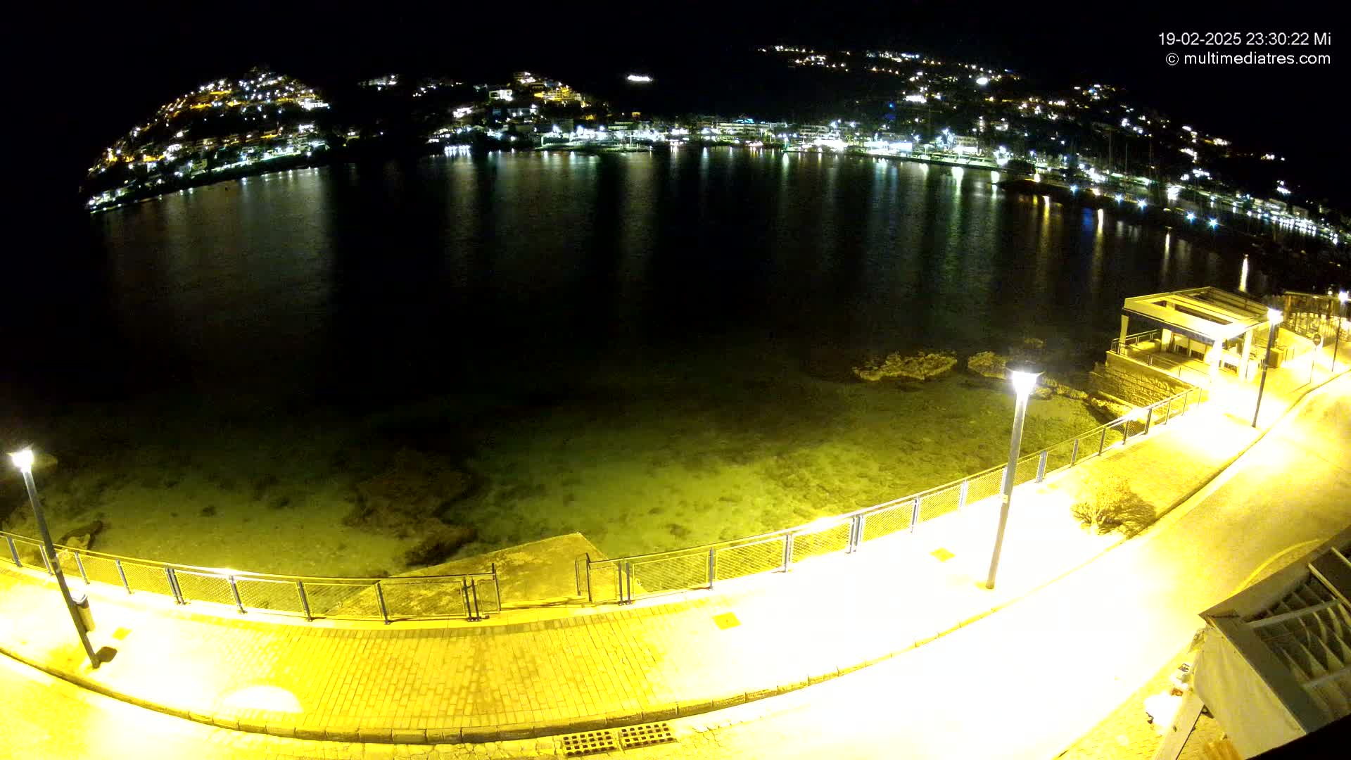 A nighttime, seaside view of a calm bay reflecting the lights of a town on a hillside, with a paved walkway and lights along the shore.