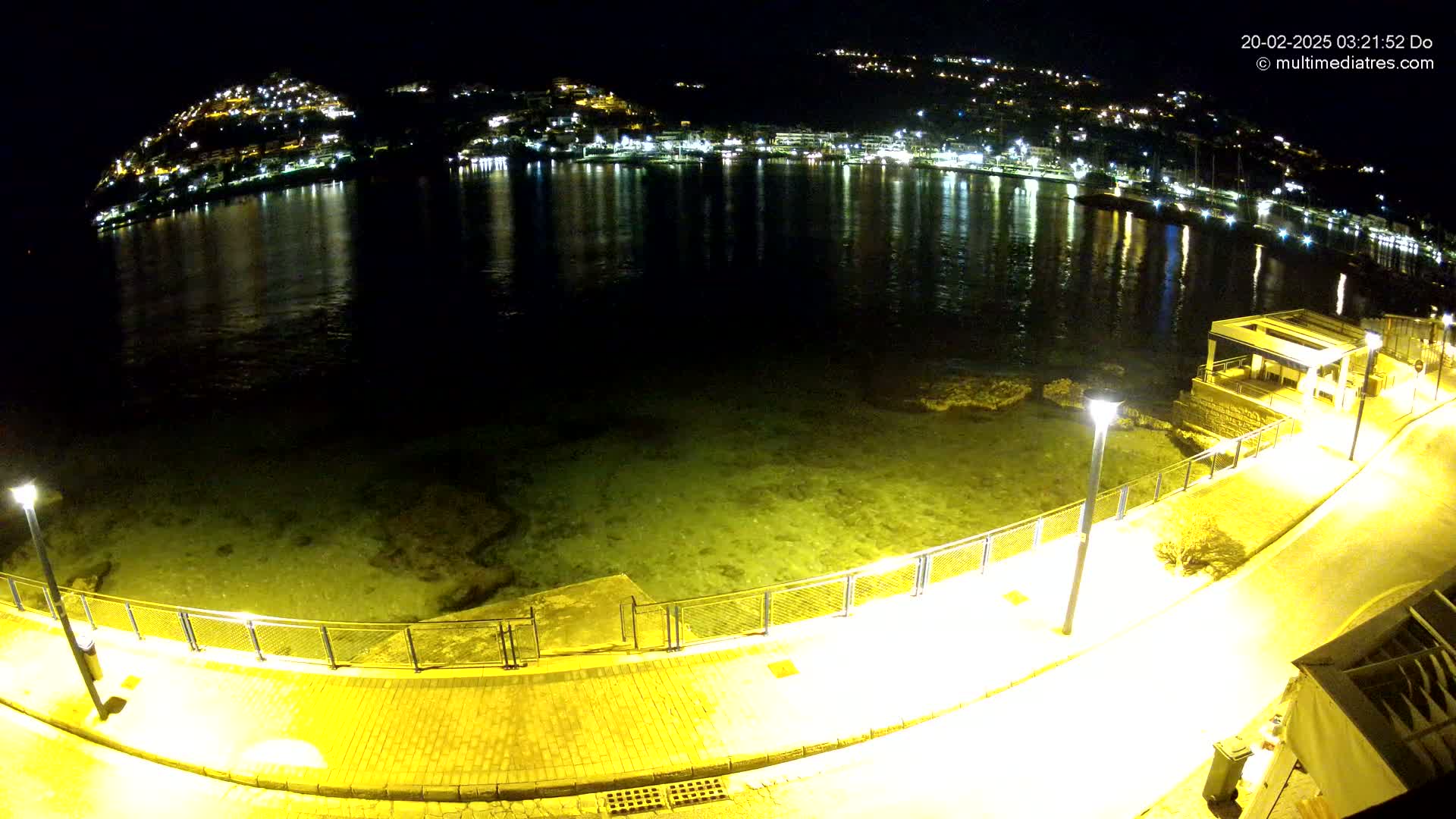 A nighttime view of a calm, shallow body of water reflecting the lights of a town built on a hillside, with a paved walkway and lampposts along the shore.