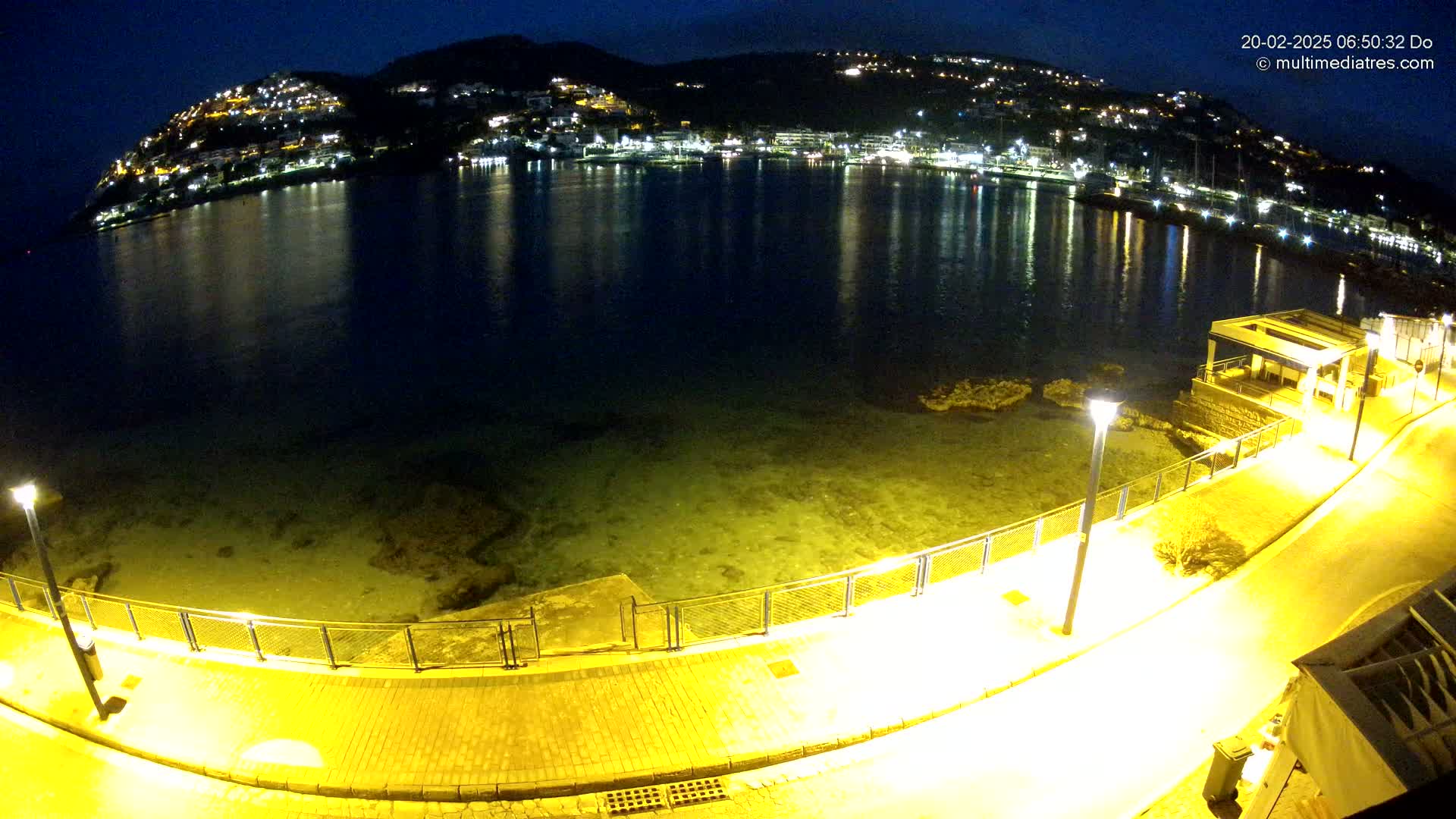A nighttime view of a calm bay, reflecting the lights of a hillside town, with a lit walkway curving along the water's edge.