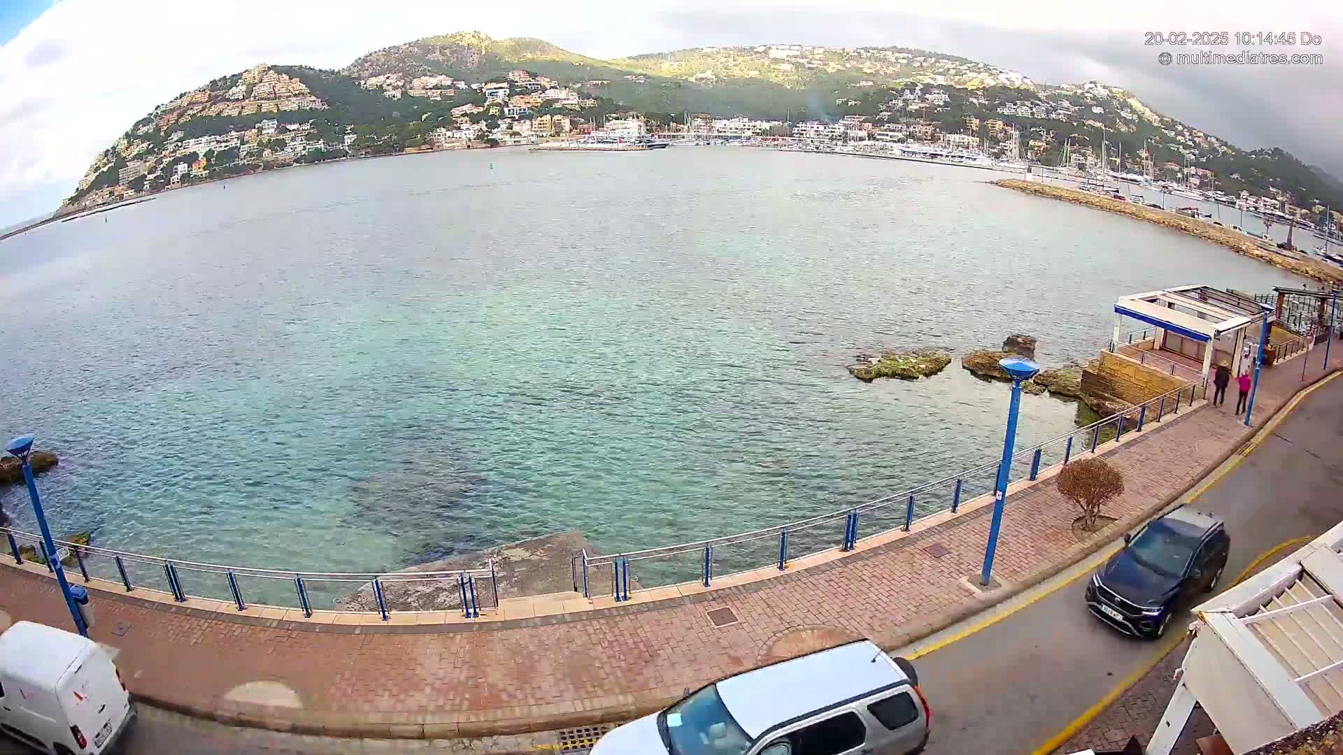 A coastal scene on an overcast day shows a calm bay with a town built on a hillside in the background, a paved walkway along the water's edge with parked cars and people walking, and a small rocky area extending into the water.