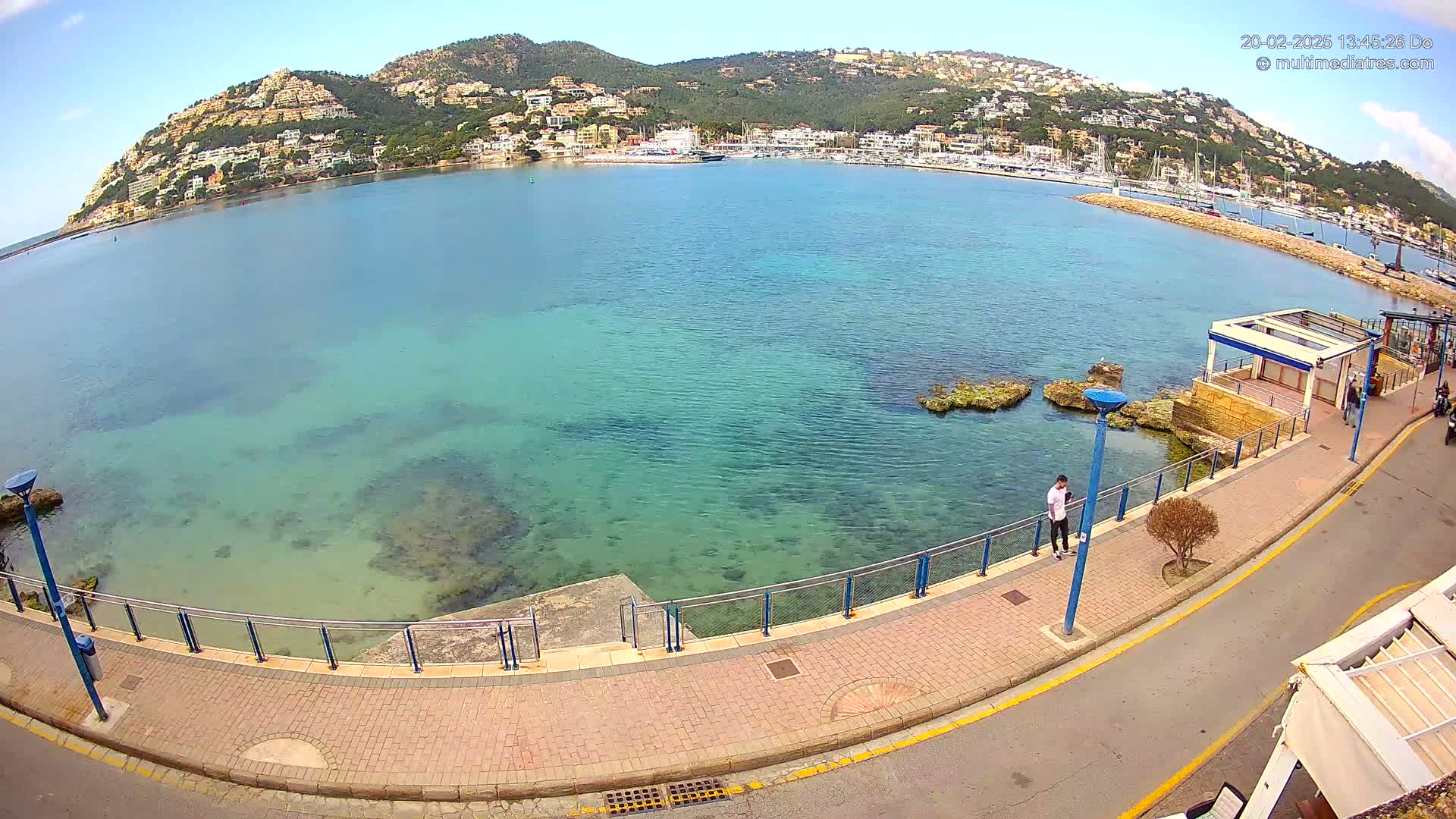A seaside promenade with a brick walkway overlooks a calm, clear turquoise bay and a hillside town under a sunny sky.