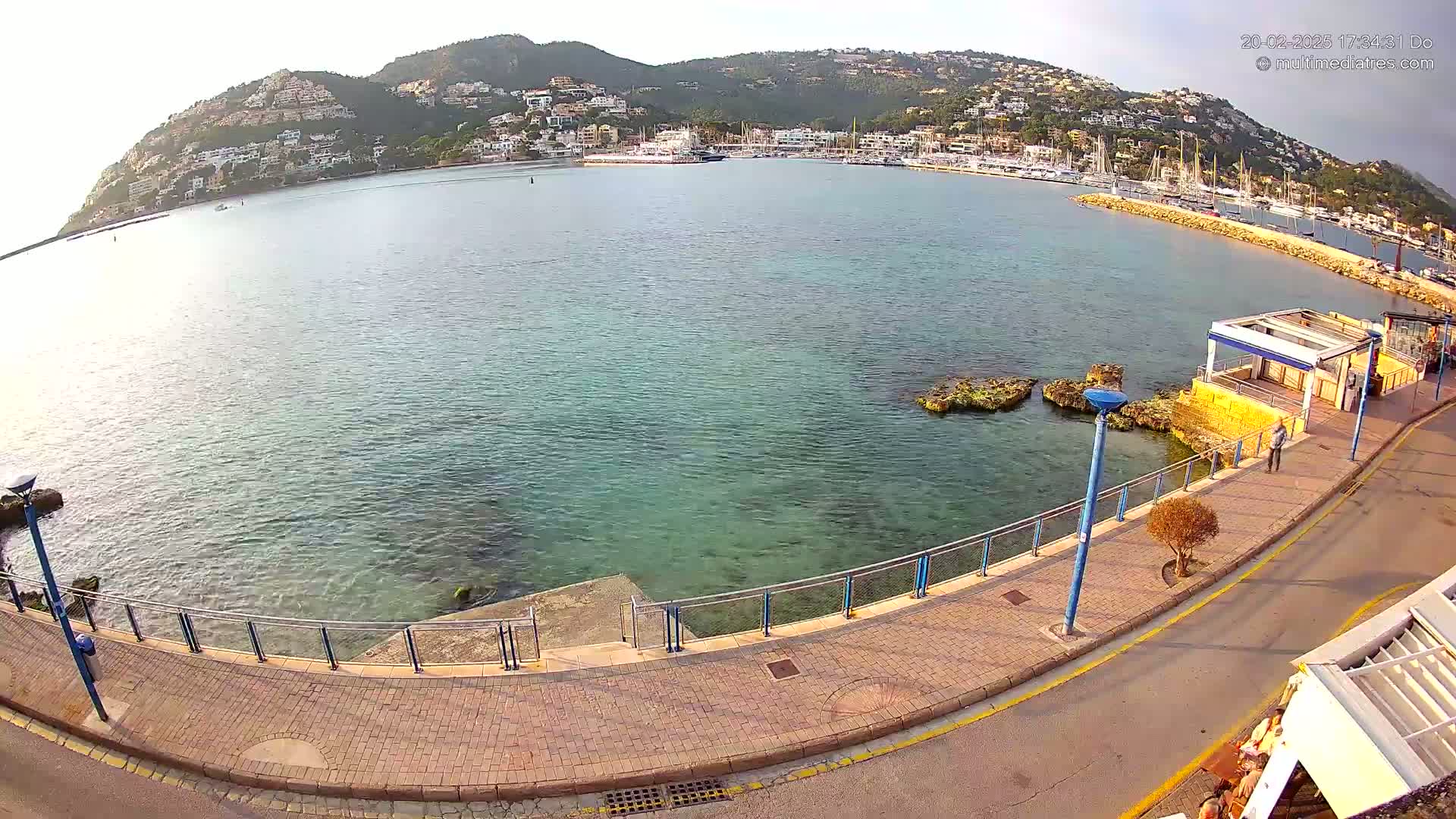 A calm, clear body of water curves around a hillside town filled with buildings and boats, all under a mostly clear sky.