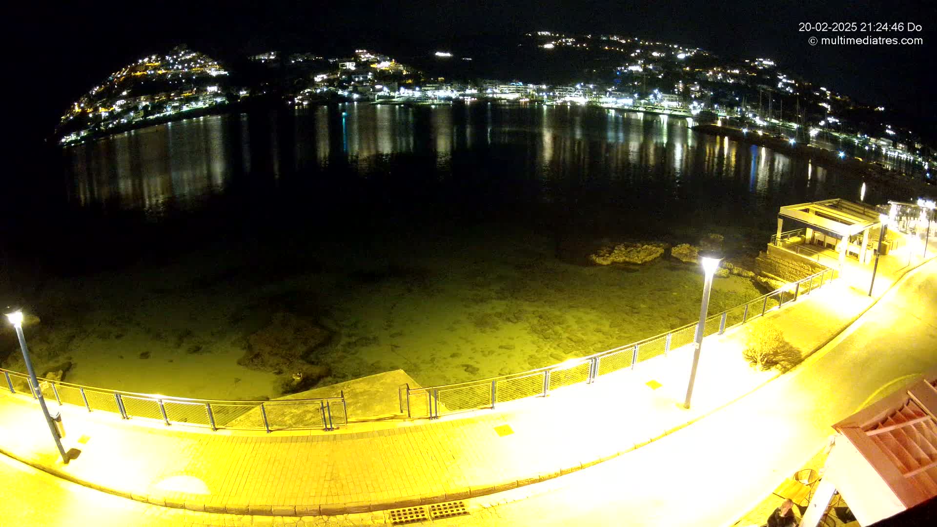 A nighttime view of a calm, lit waterfront with a paved walkway, low seawall, and a hilltop town brightly illuminated across the water.