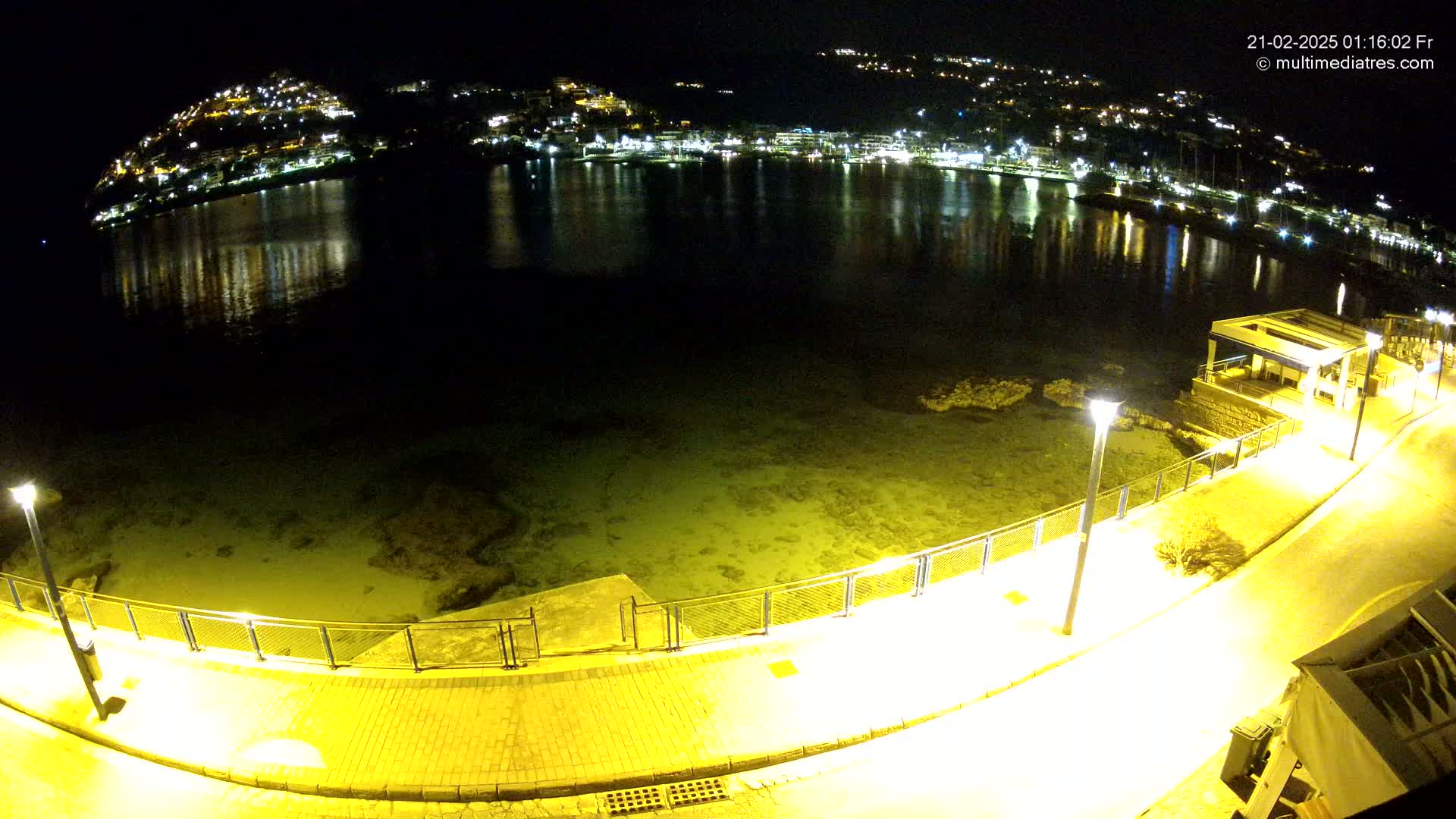 A nighttime, high-angle view shows a calm body of water reflecting the lights of a town built on a hillside, with a paved walkway and lights illuminating the shoreline.