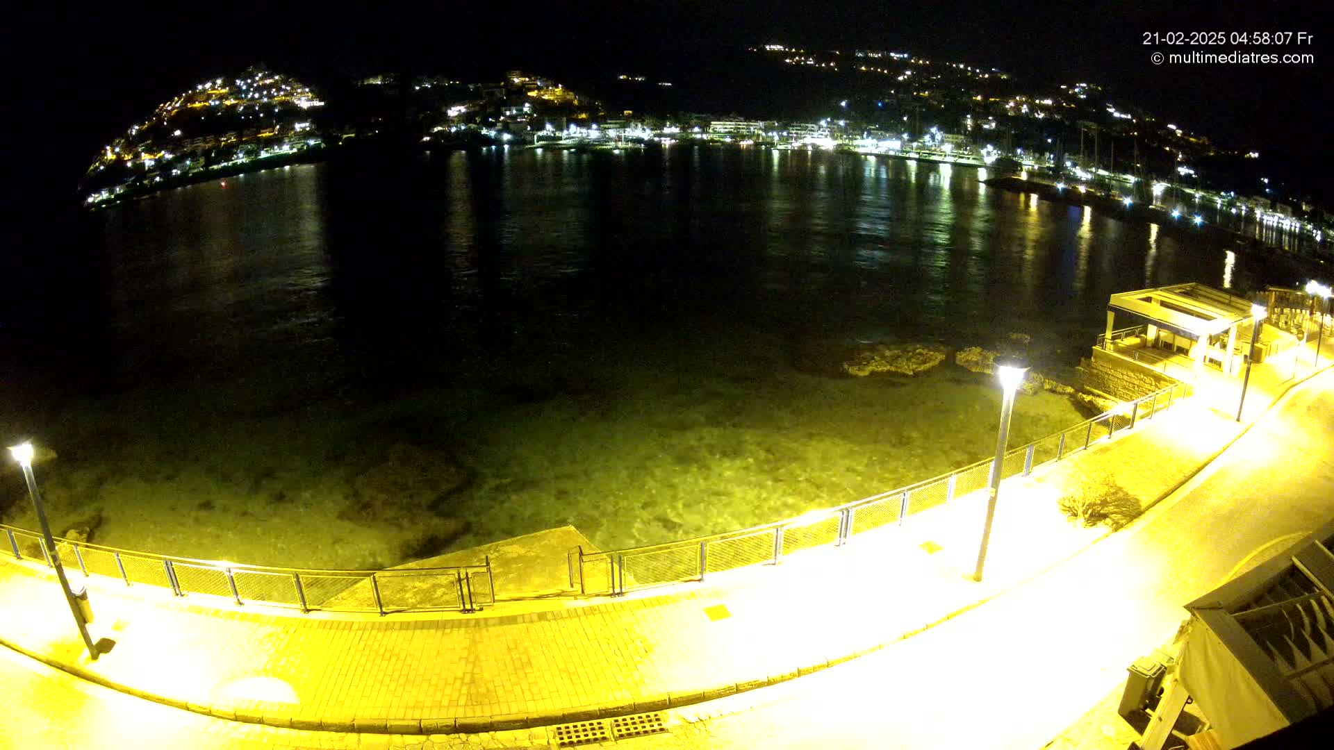 A nighttime, high-angle view shows a paved walkway along a calm body of water, illuminated by streetlights, with a hilltop town shimmering with lights in the background.