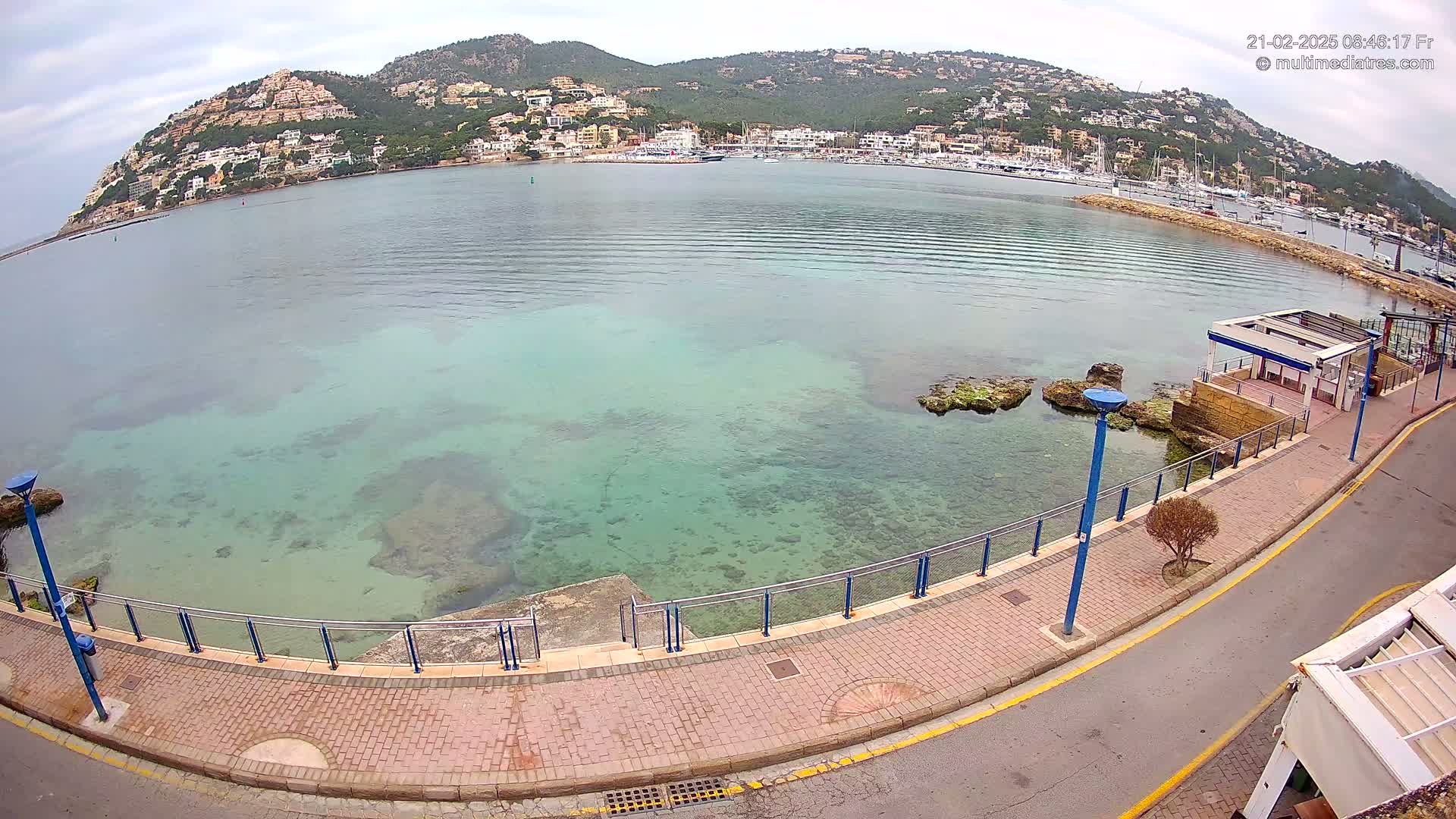 A paved walkway overlooks a calm, clear, teal-colored bay with a town and hills in the background under a cloudy sky.