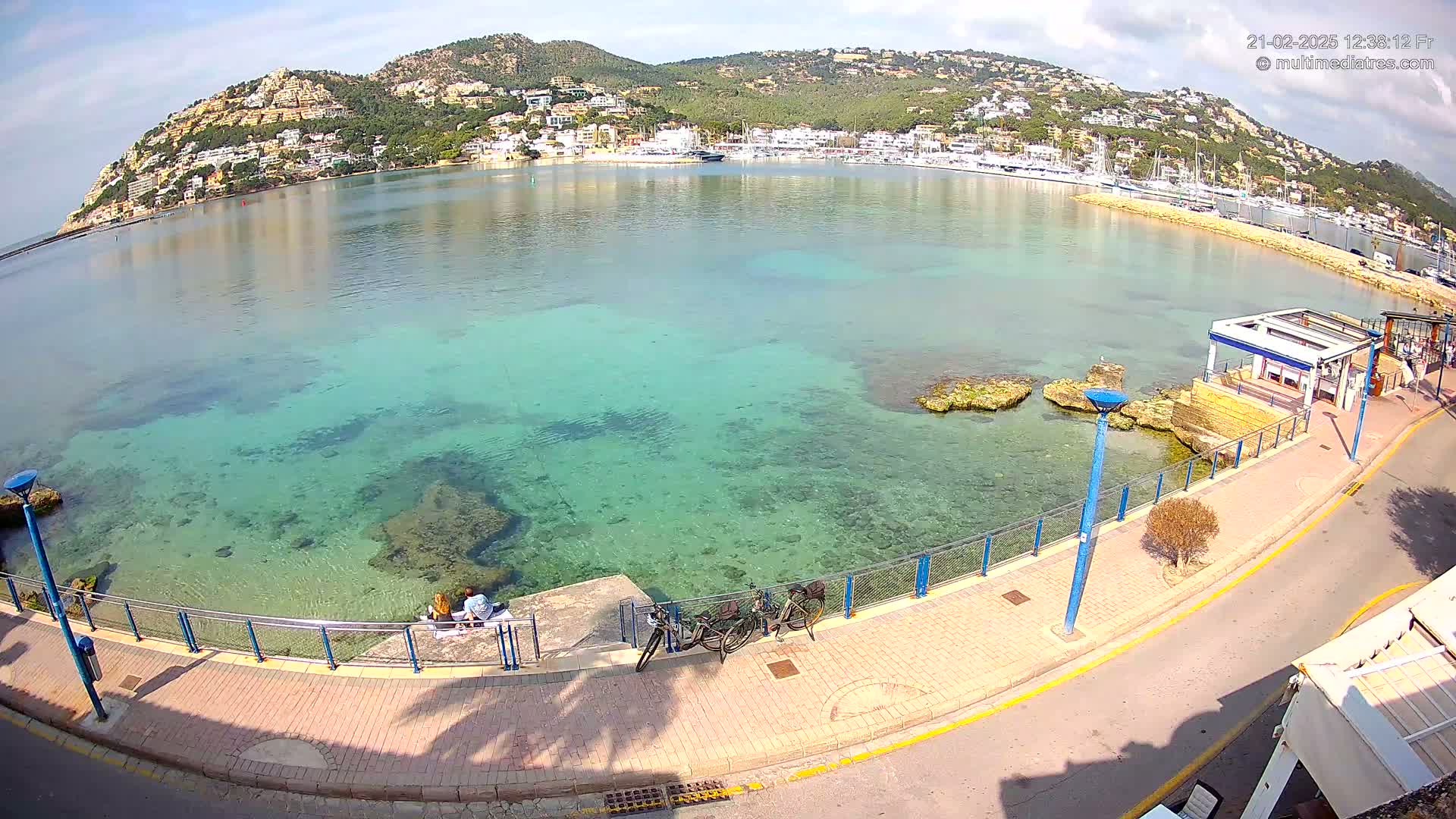 A clear, sunny day shows a calm, shallow, teal-colored bay with a town and hills in the background, viewed from a waterfront promenade.