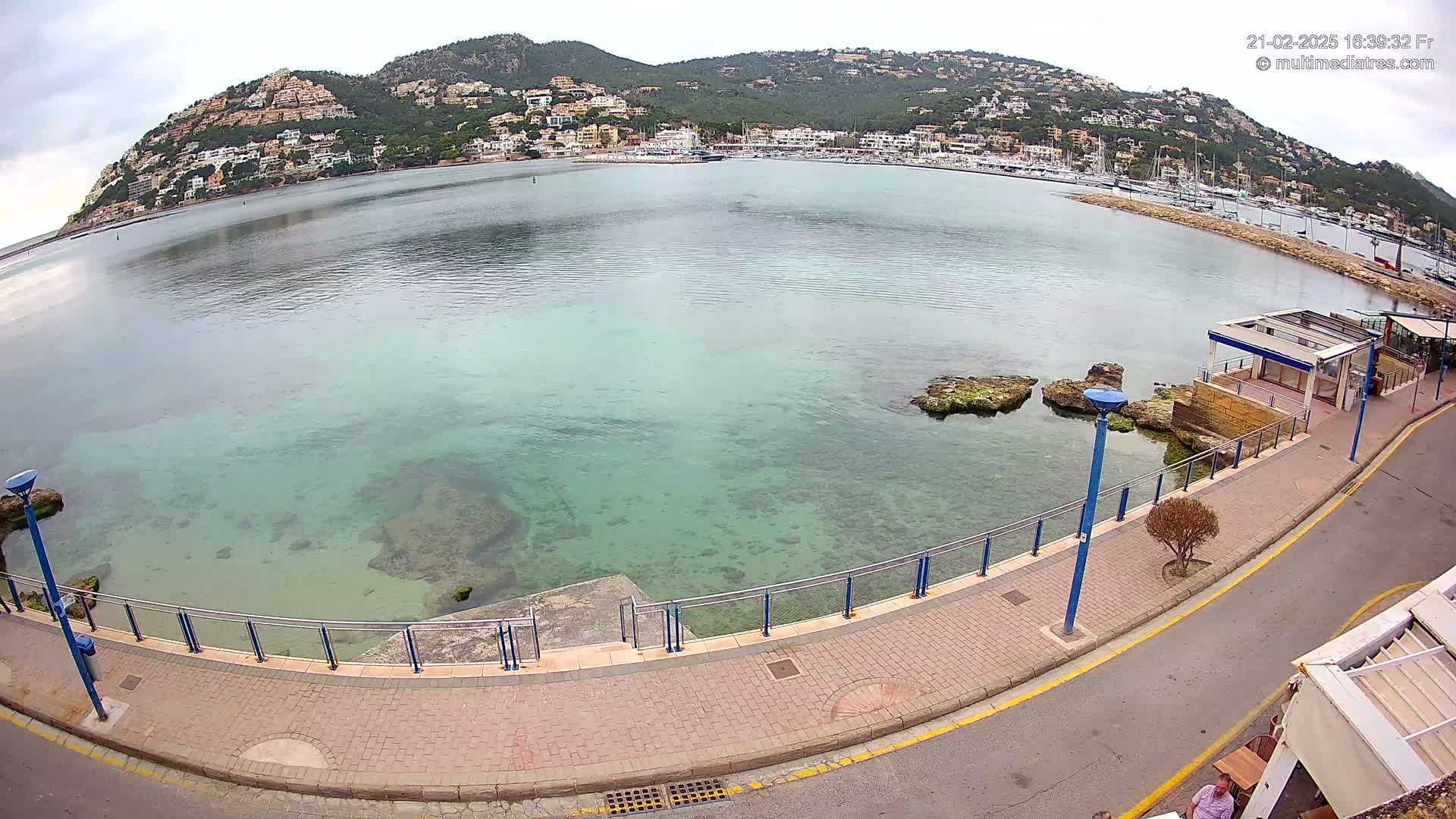 A paved walkway with a railing overlooks a calm, clear bay with a town nestled against a hillside in the background under an overcast sky.