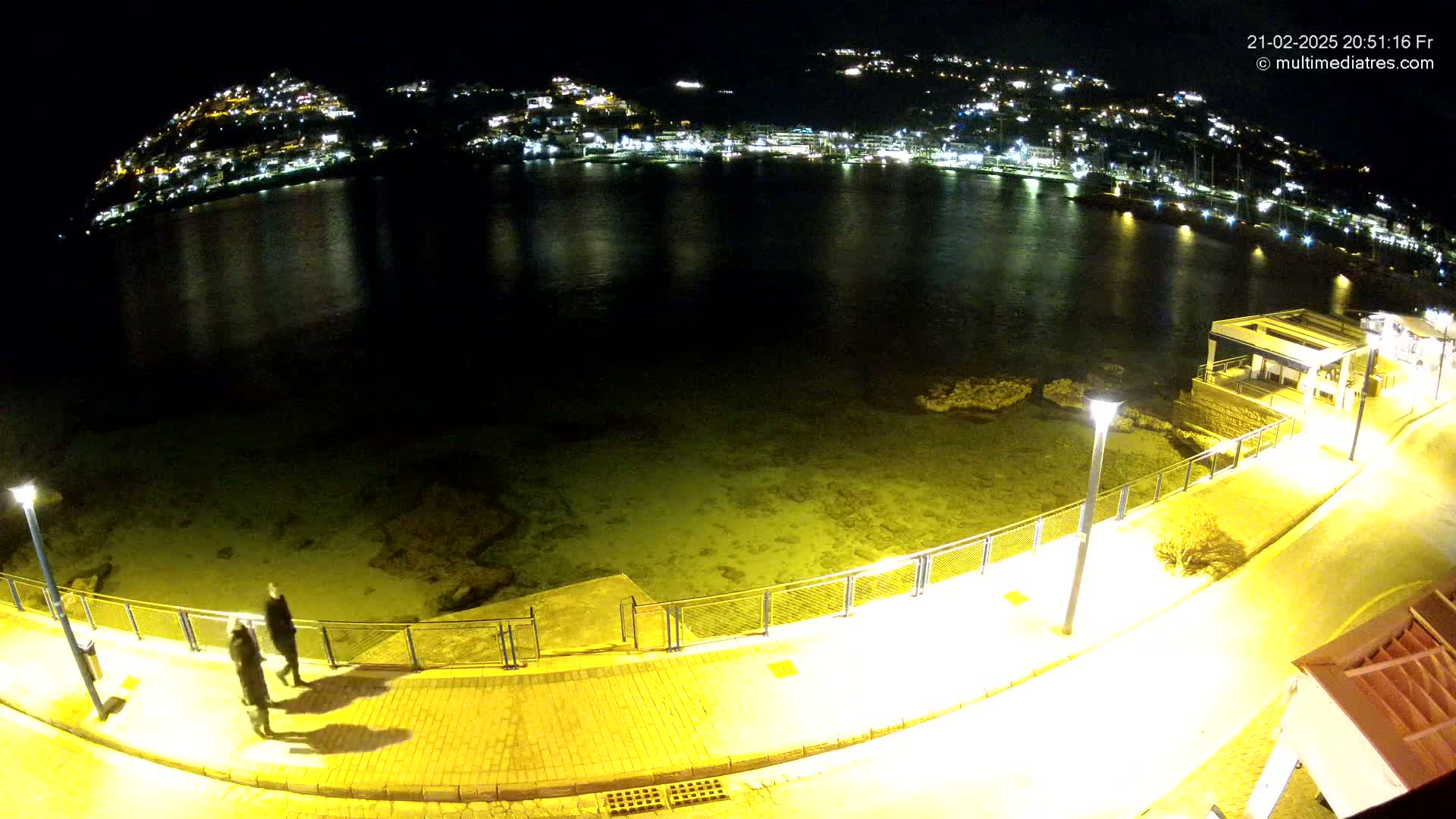 A nighttime, high-angle view shows a calm body of water reflecting the lights of a town on a hillside, with a paved walkway and two people walking along the waterfront.