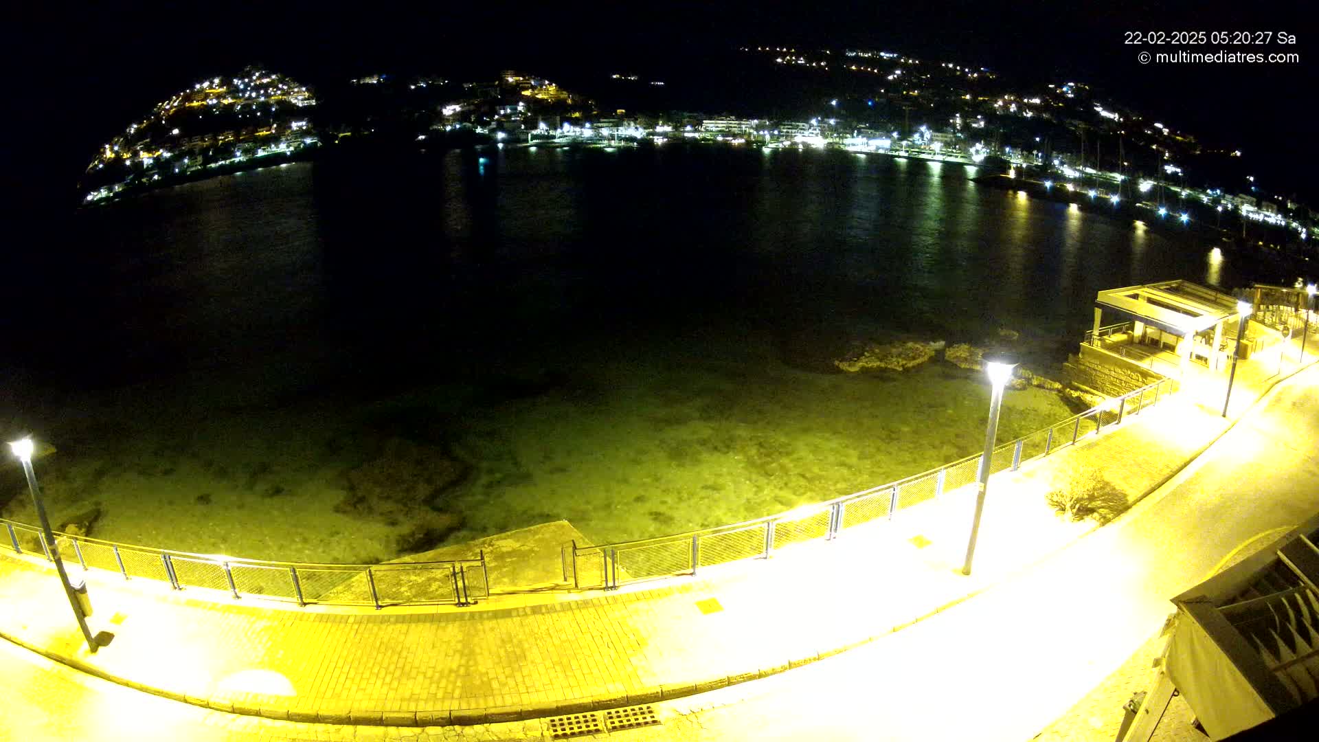 A nighttime view of a calm bay with a lit waterfront path and a hill dotted with lights in the background.