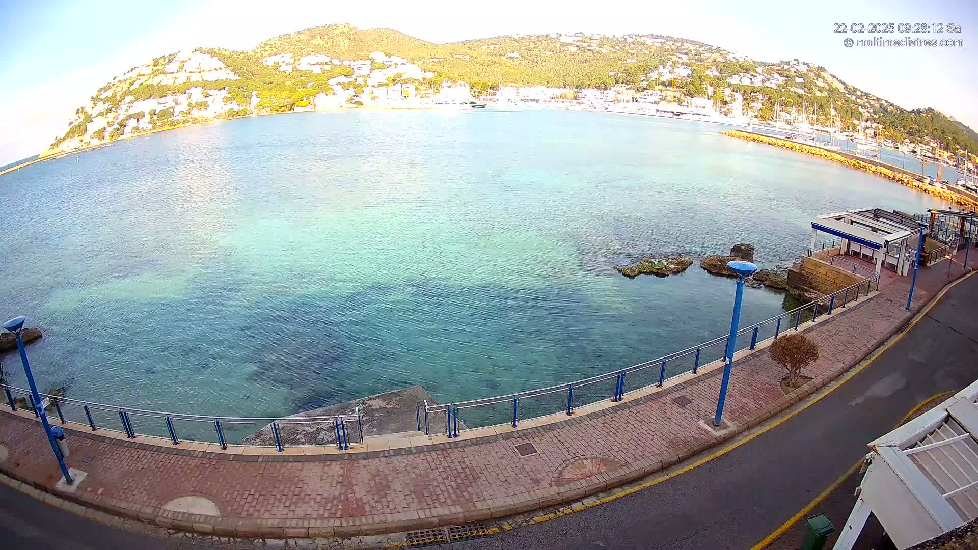 A clear, sunny day overlooks a calm, teal-colored bay with a town nestled on a hill in the background, bordered by a paved walkway and a small road.