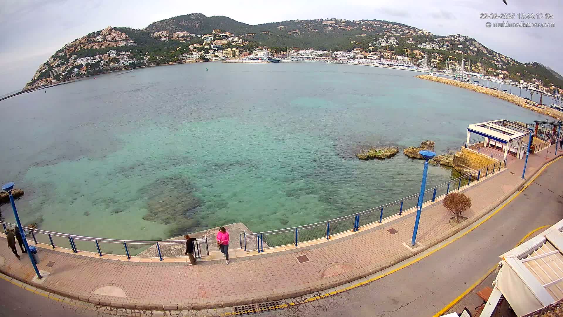 A seaside promenade overlooks a calm, clear turquoise bay with a hilltop town and a marina in the background under an overcast sky.