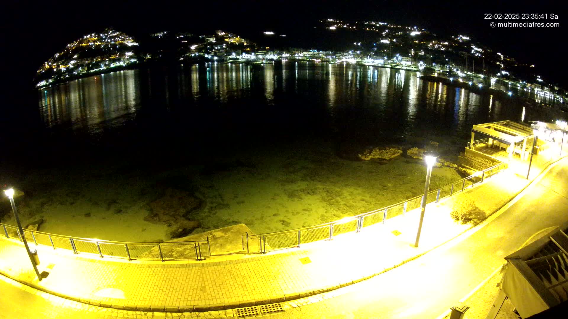 A nighttime view of a waterfront promenade illuminated by lampposts, overlooking a calm bay reflecting the lights of a hillside town.