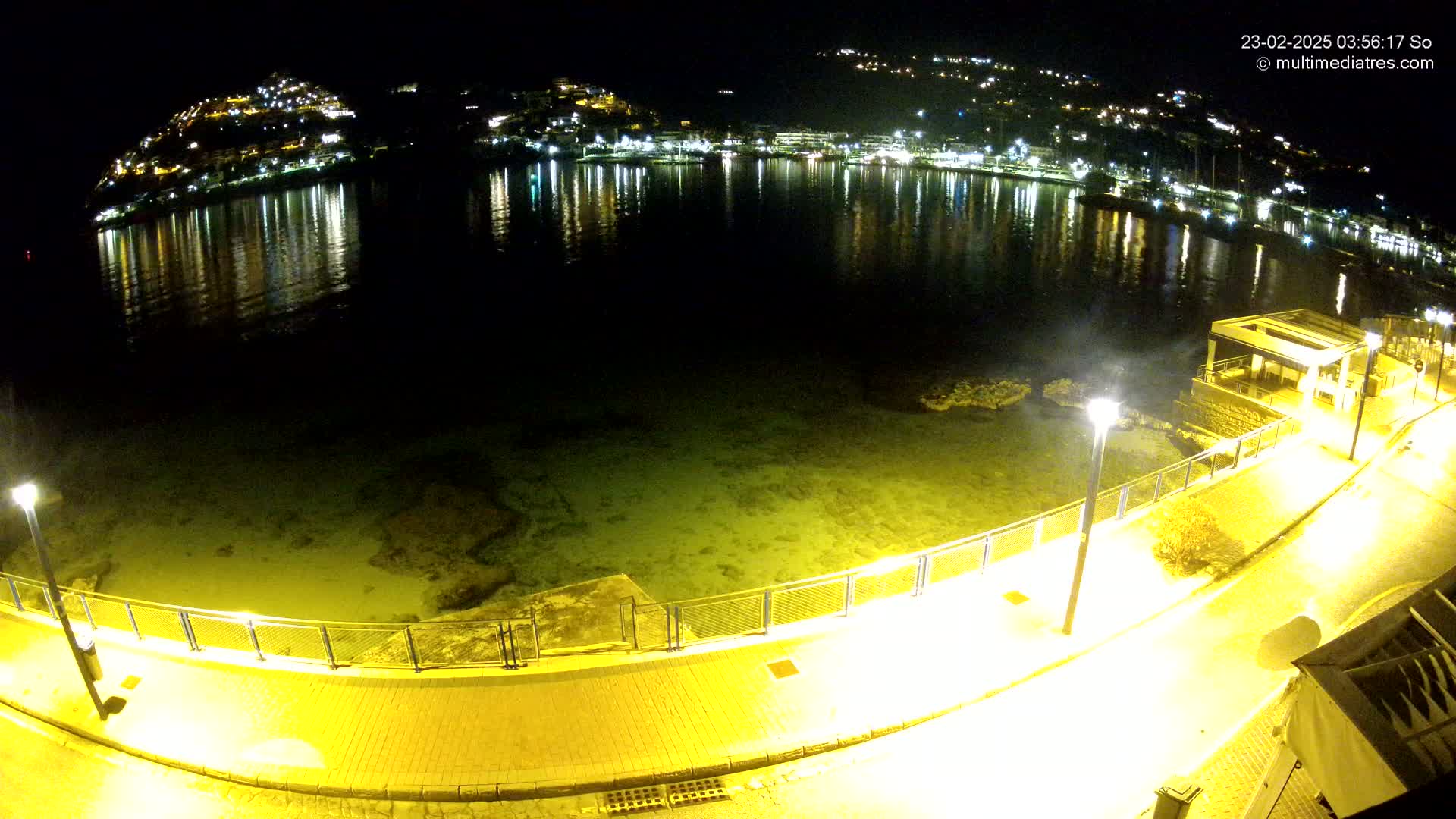 A nighttime view of a calm, shallow body of water reflecting the lights of a town on a hillside, with a brightly lit paved walkway and railing along the shore.