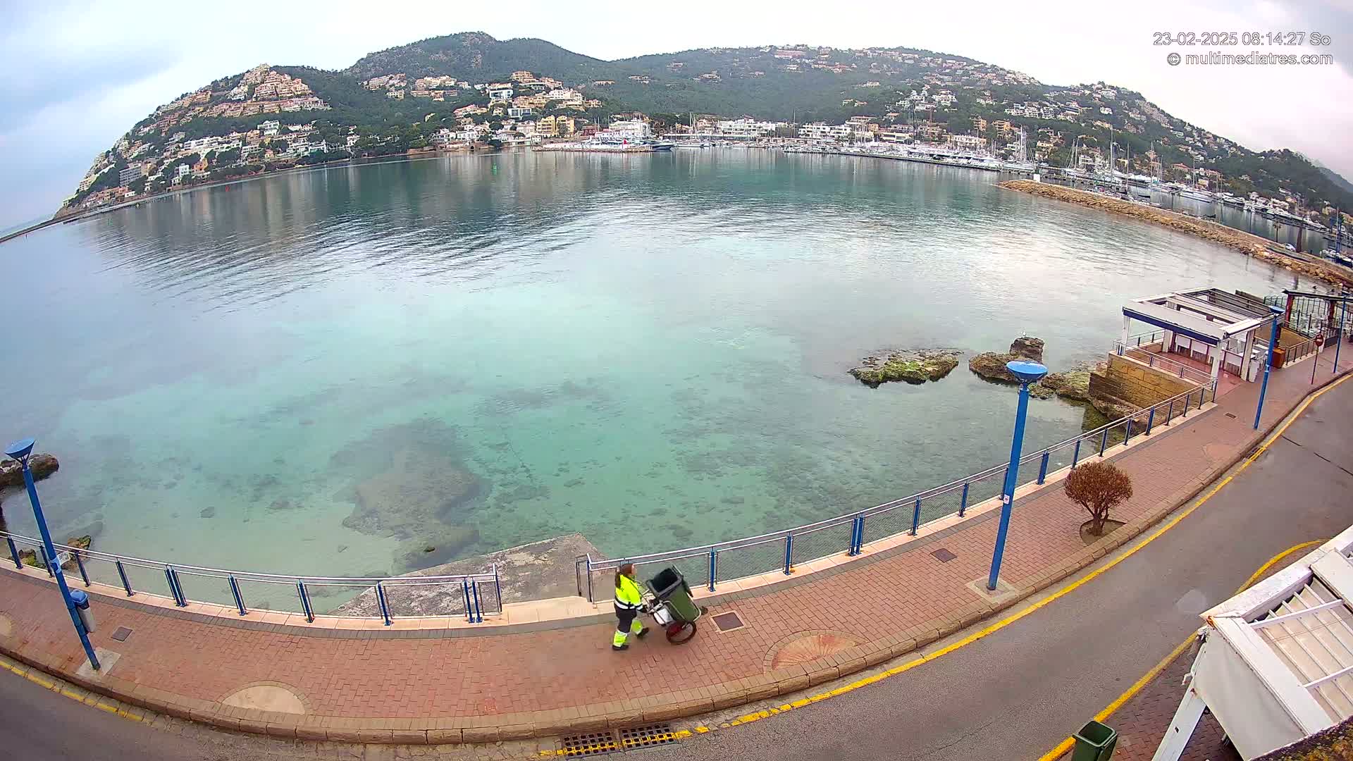 A person in a bright green jacket pushes a cart along a waterfront walkway overlooking a calm, clear bay with a hill of buildings in the background on an overcast day.