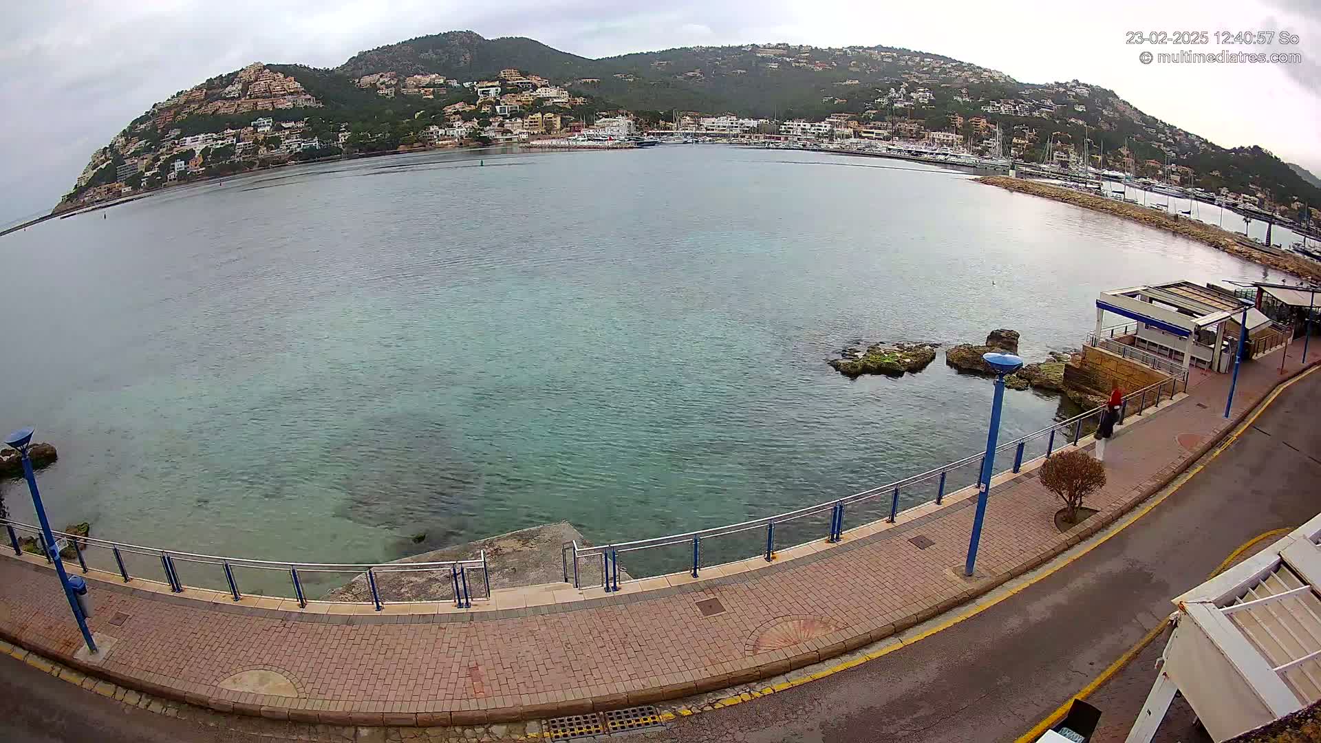 A seaside promenade with a brick walkway and metal railing overlooks a calm, greenish-blue bay, with a hill dotted with buildings in the background under an overcast sky.