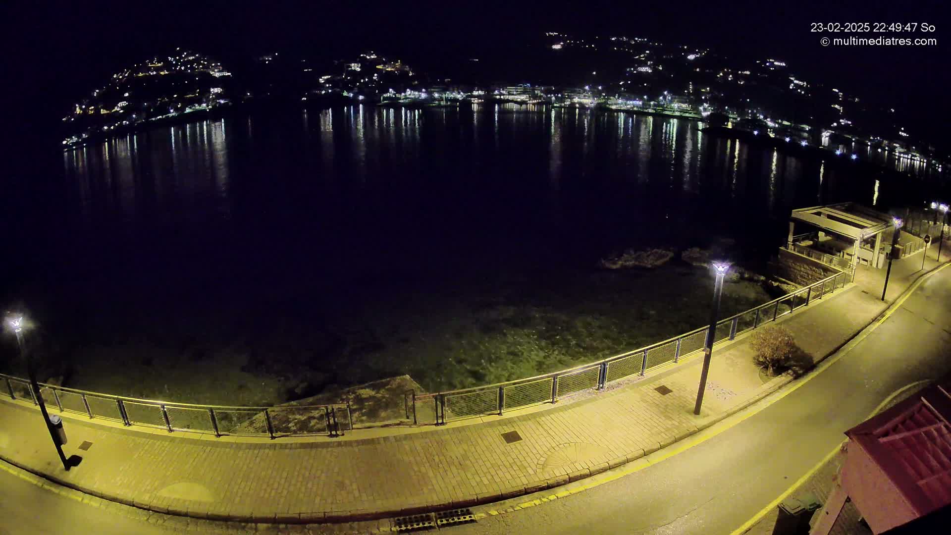 A nighttime view of a calm body of water reflecting the lights of a town on the opposite shore, with a paved walkway and lampposts along the waterfront.