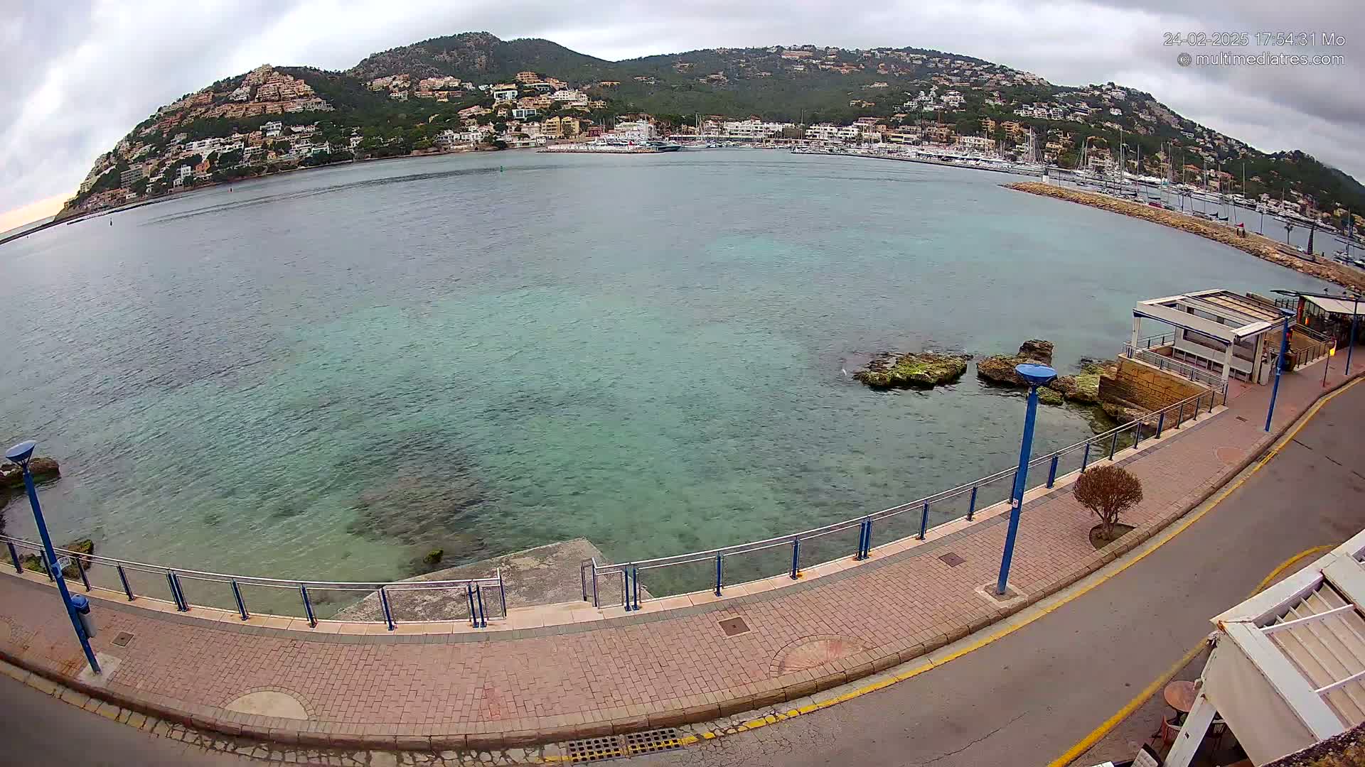 A paved walkway curves along a calm, clear, teal-colored body of water, overlooking a small harbor filled with boats and a hillside town under a cloudy sky.