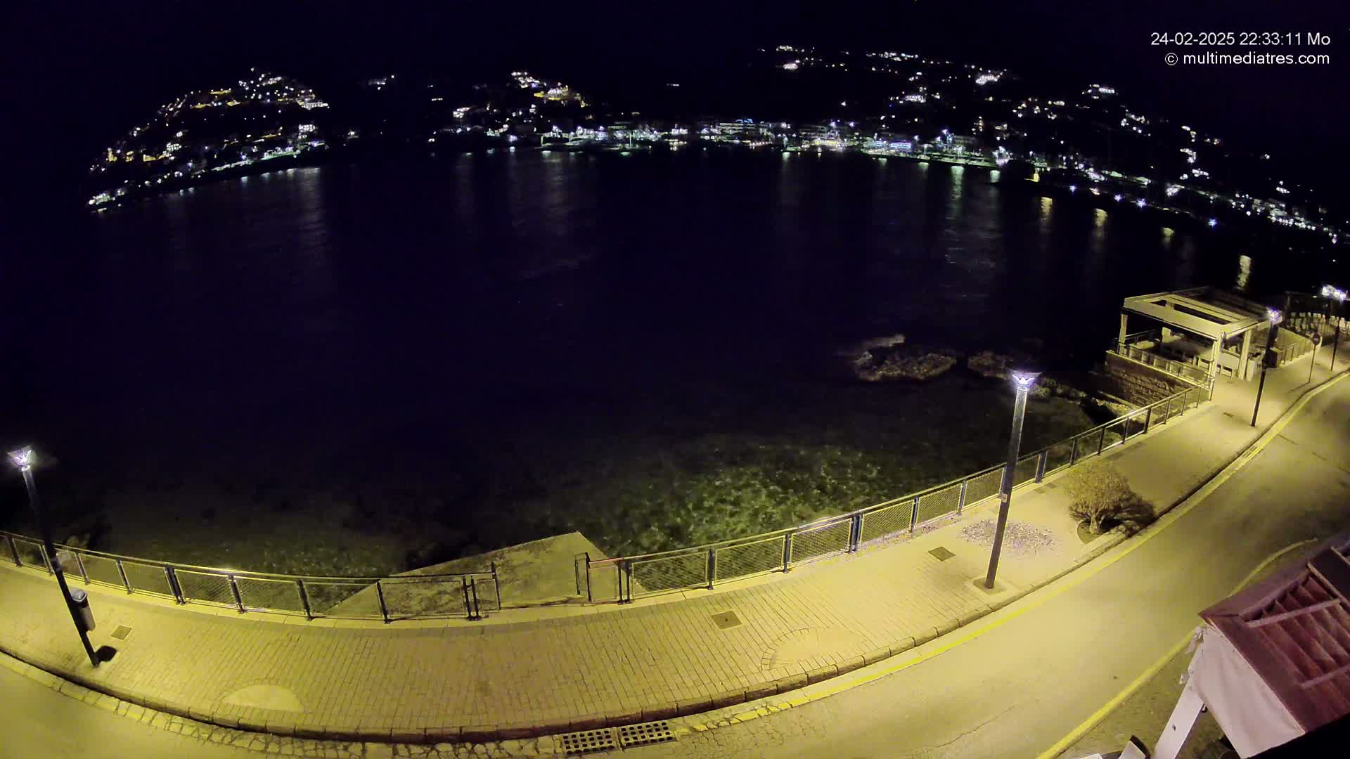 A nighttime view of a paved walkway beside a calm body of water, with a hill of illuminated buildings in the background.