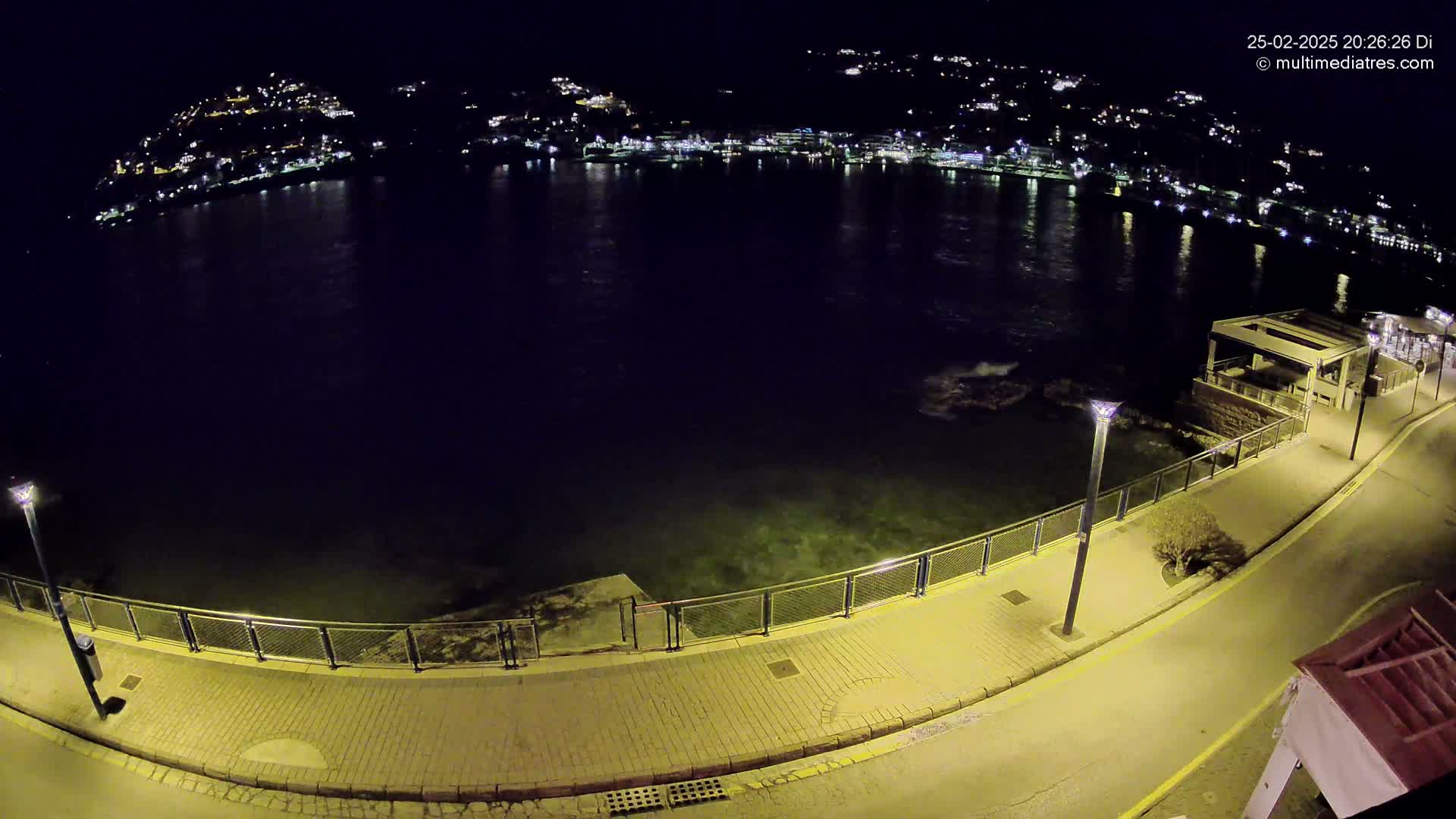 A nighttime, high-angle view shows a paved walkway next to a calm body of water, with a town's lights visible across the water in the distance.
