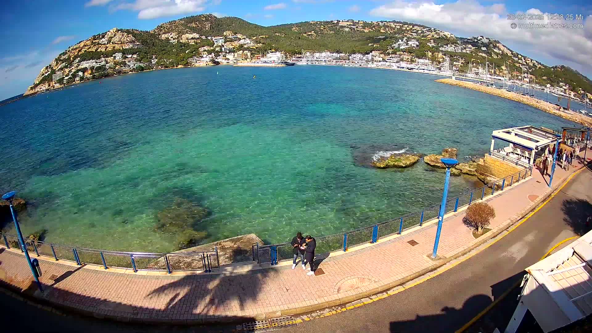 A clear, sunny day reveals a picturesque waterfront scene with calm, teal water, a paved walkway, and a hill dotted with buildings in the background.