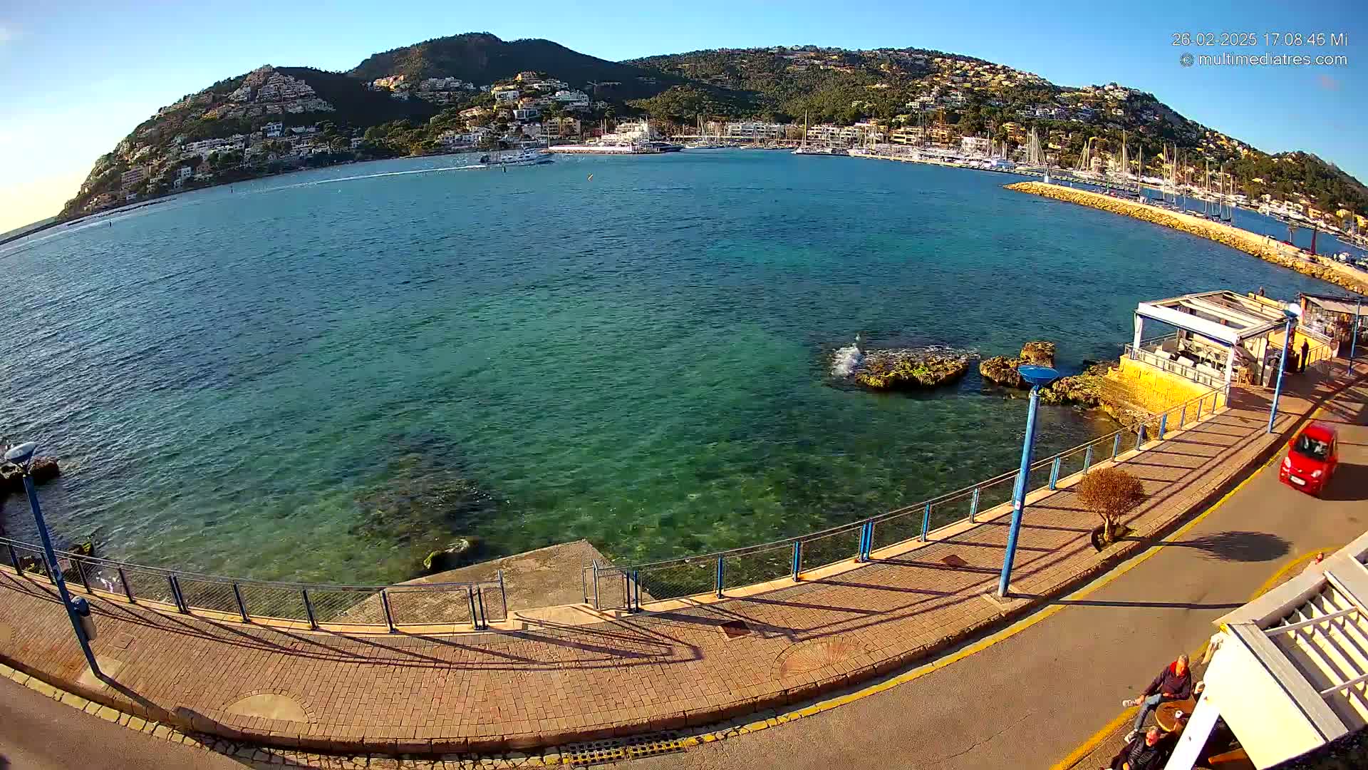 A clear, sunny day reveals a seaside promenade overlooking a calm, teal-colored bay with a hilltop town in the background.
