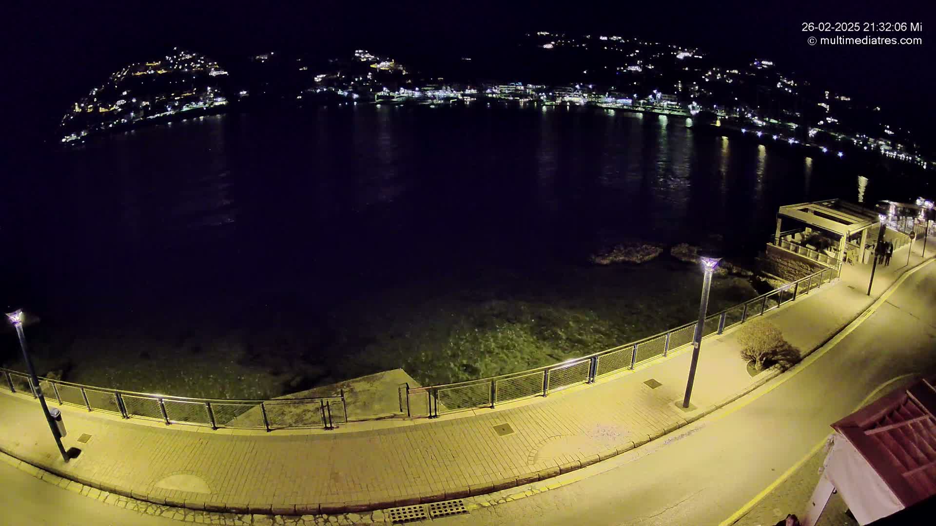 A nighttime, high-angle view shows a paved walkway along a calm body of water, with a hill of illuminated buildings in the background.