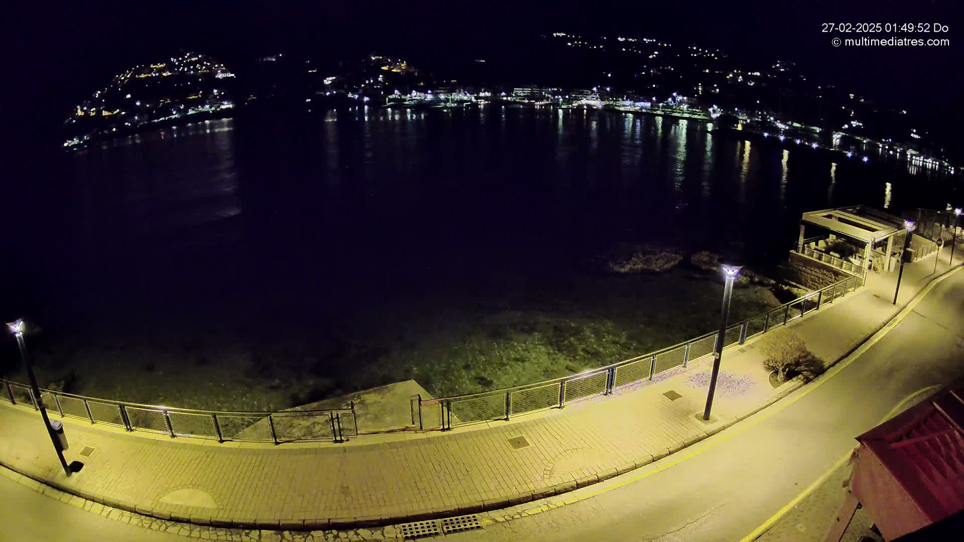A nighttime view of a paved waterfront walkway with lights, overlooking a calm, dark body of water reflecting the lights of a town on the opposite shore.