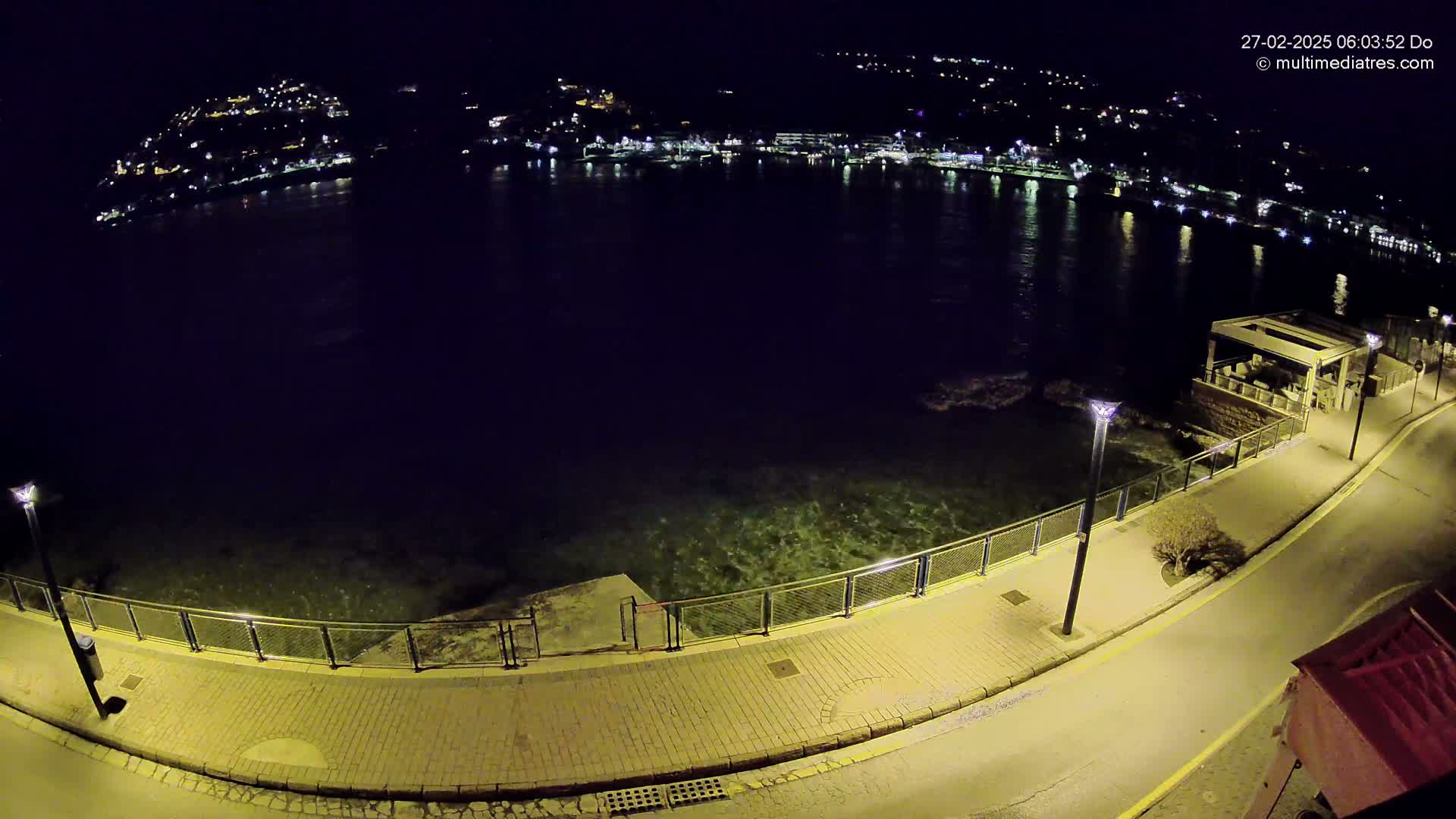 A nighttime, high-angle view shows a paved waterfront path next to calm dark water, with a distant town illuminated by lights across the water.
