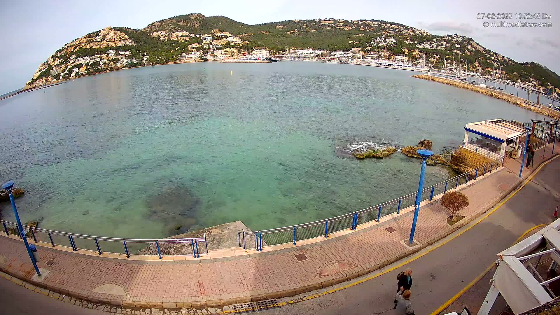 A paved walkway curves along a calm, clear teal sea, with a small building and a hill covered in buildings in the background under a mostly clear sky.