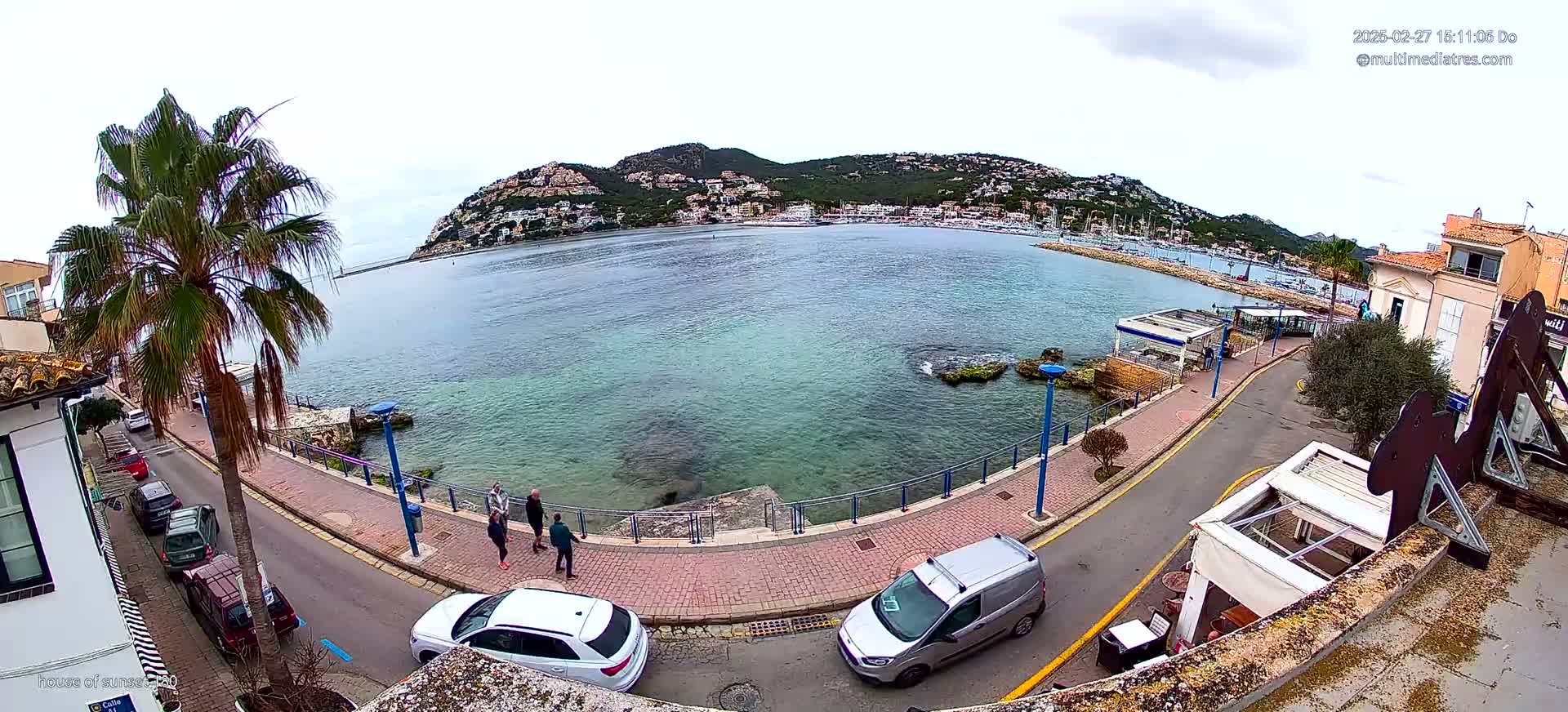 A coastal town scene on an overcast day shows a calm bay with a hill covered in buildings in the background, a paved walkway along the water's edge, several parked cars, and a few pedestrians.