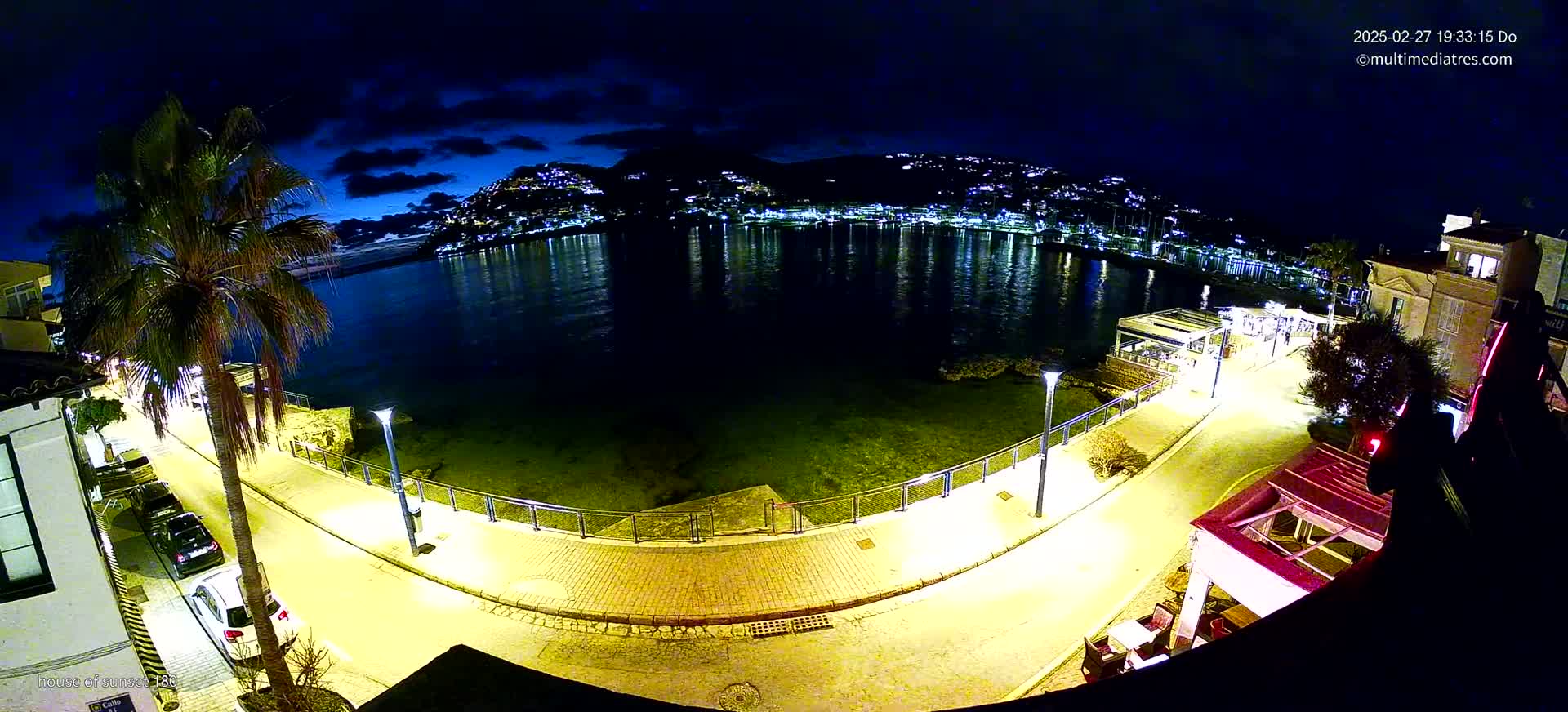 A nighttime, high-angle view of a waterfront promenade illuminated by streetlights, with a calm body of water reflecting the lights of buildings on the opposite shore.