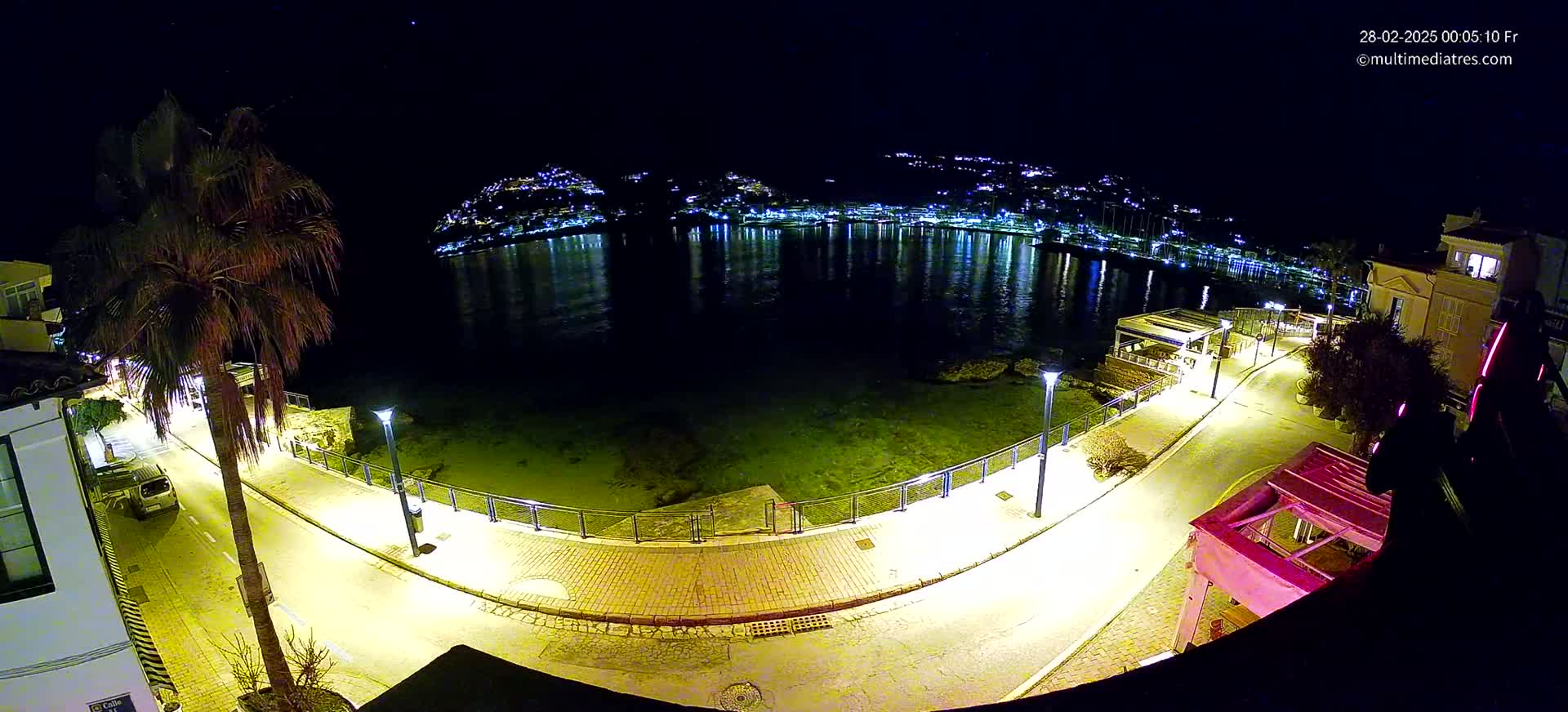 A nighttime aerial view shows a calm body of water reflecting the lights of a town on the opposite shore, with a palm tree and a paved walkway illuminated along the waterfront.
